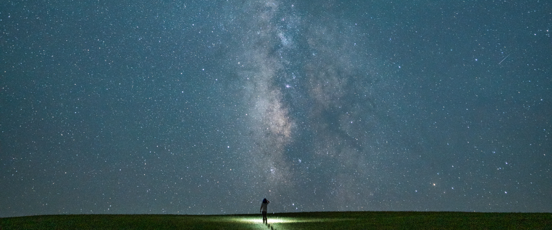 A person stands on a grassy hill at night, illuminated by a flashlight, gazing up at the Milky Way galaxy and a sky filled with countless stars.