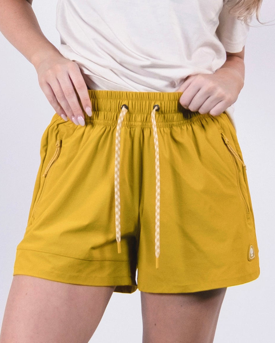 A person stands against a light background with hands on hips, wearing a white T-shirt, BEARDEDGOAT Women's Alpina Short in mustard yellow with zippered pockets, plus white socks and sneakers.