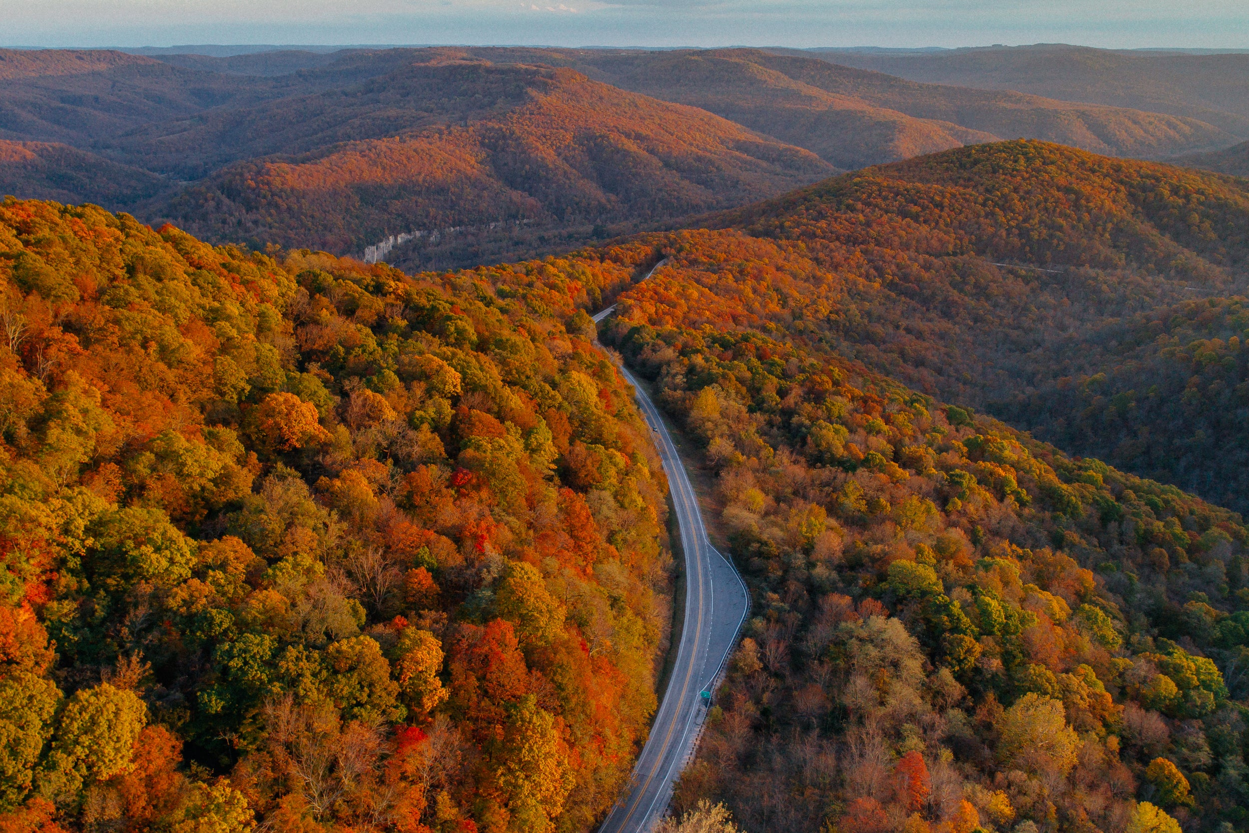 A winding road cuts through rolling hills covered in autumn foliage, with vibrant red, orange, yellow, and green trees stretching into the distance under a clear sky.