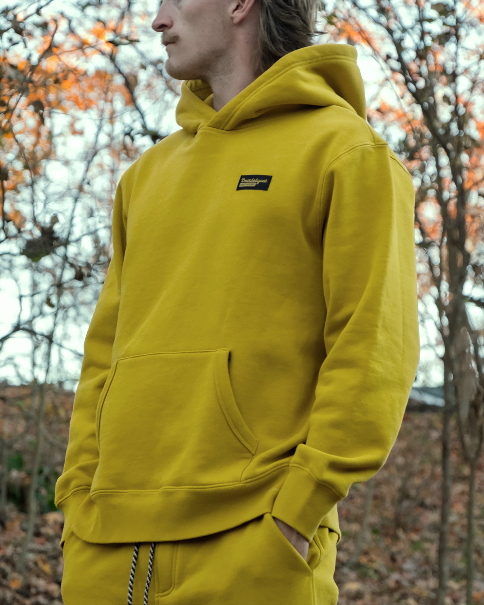 A person stands outdoors among autumn leaves and bare trees, facing sideways with hands in their pocket, wearing the bright yellow beardedgoat The VOID™ Hoodie made from heavyweight cotton fleece.