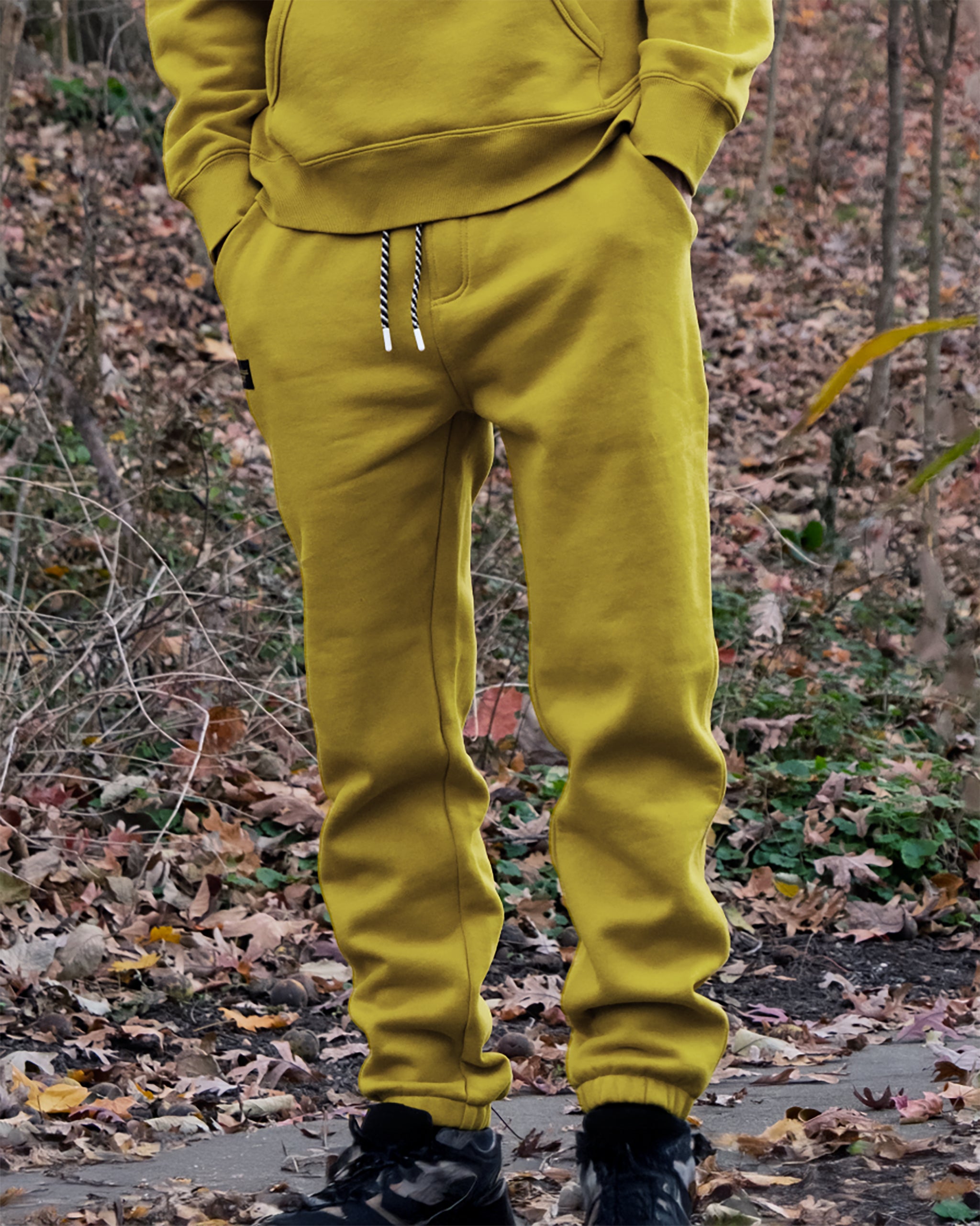 Someone wearing beardedgoat VOID™ Sweatpant and a mustard yellow hoodie with black shoes stands outdoors on a path covered in fallen autumn leaves, their face not visible.