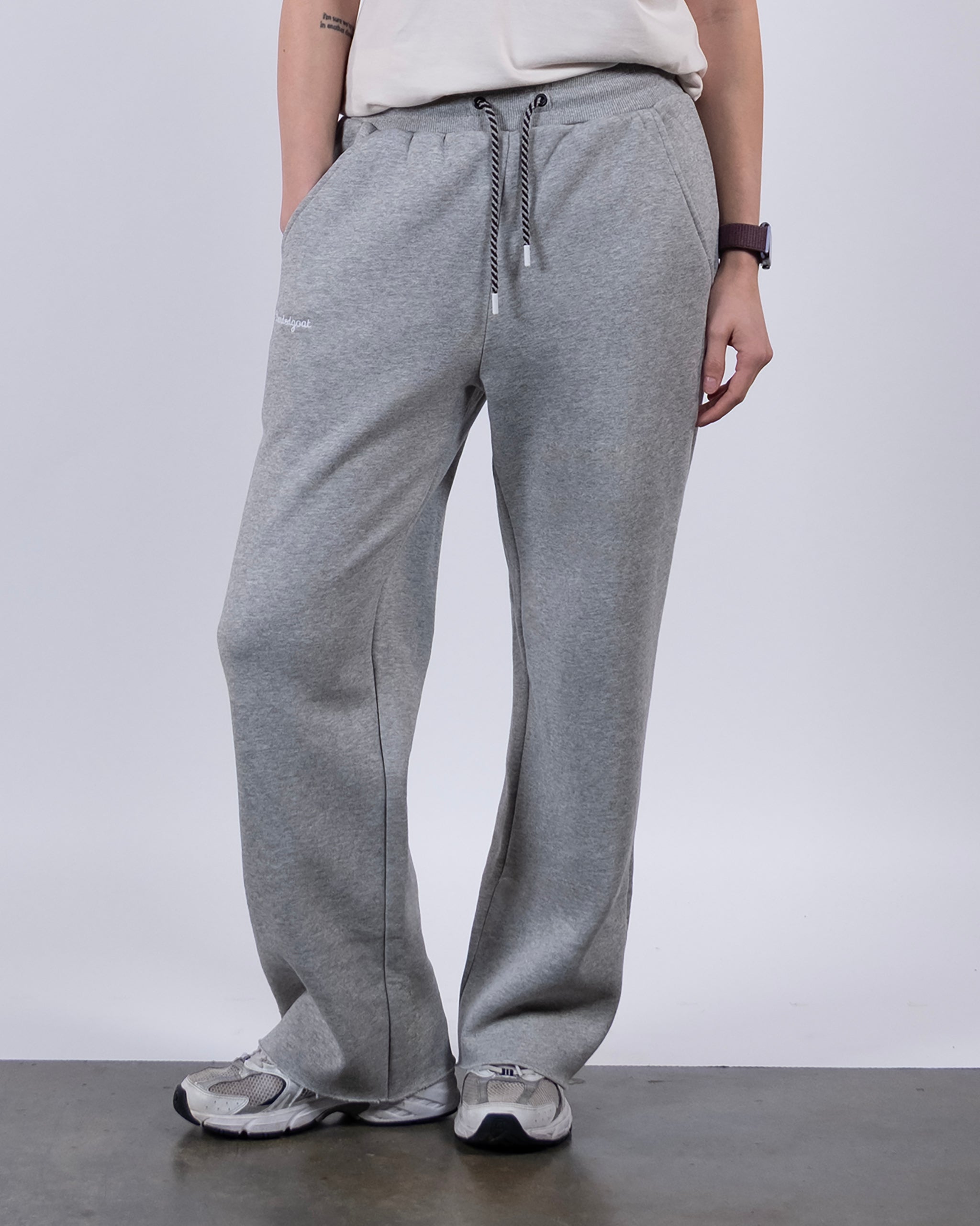 A person in beardedgoat Women’s Overnight Sweatpant in gray, a white tee, white sneakers, and a purple watch stands on a gray floor with hands in pockets. The image is cropped at the shoulders.