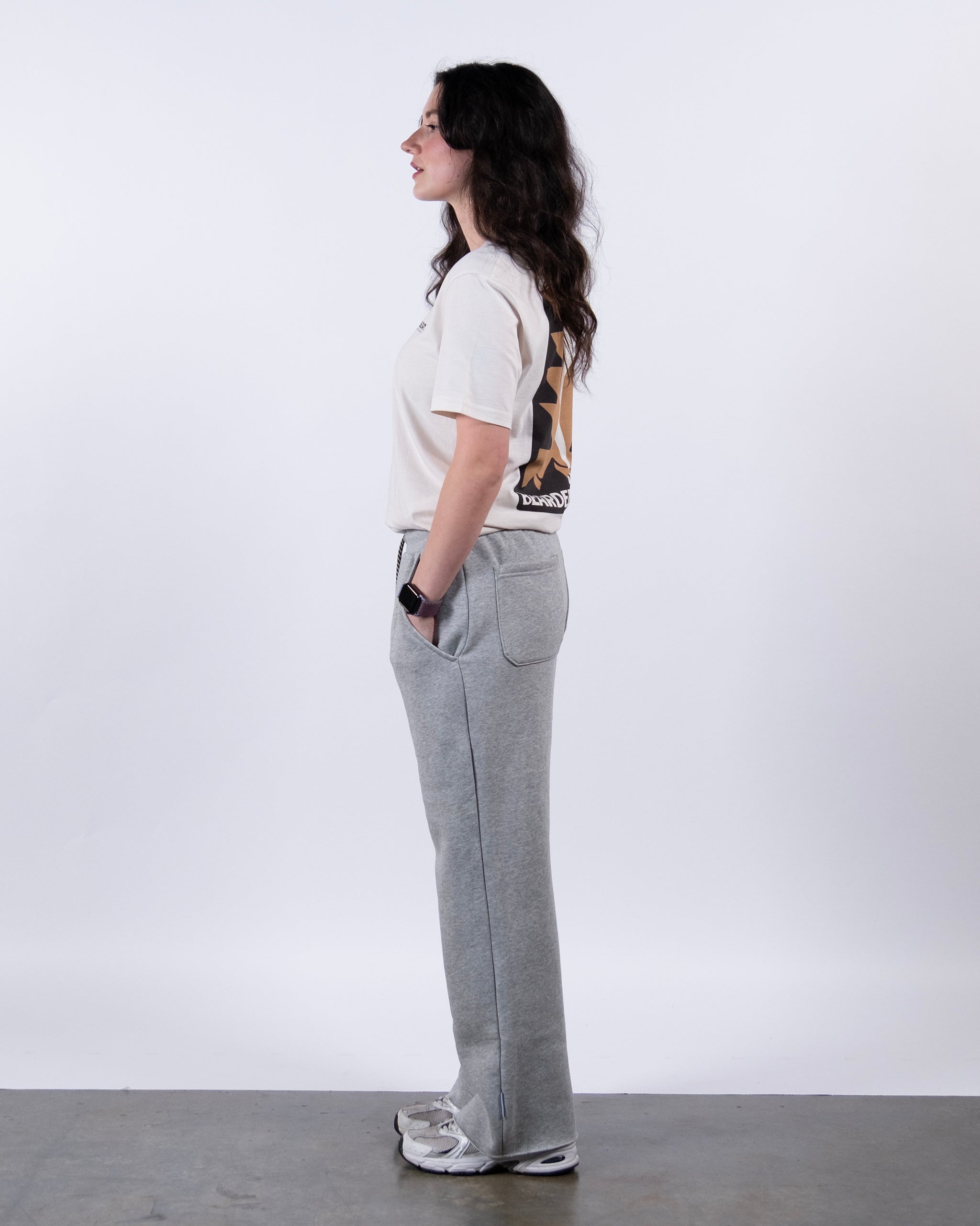 A woman with long dark hair stands in profile against a white background, wearing a light T-shirt, gray beardedgoat Women’s Overnight Sweatpant, and sneakers, with hands tucked casually into her pockets.