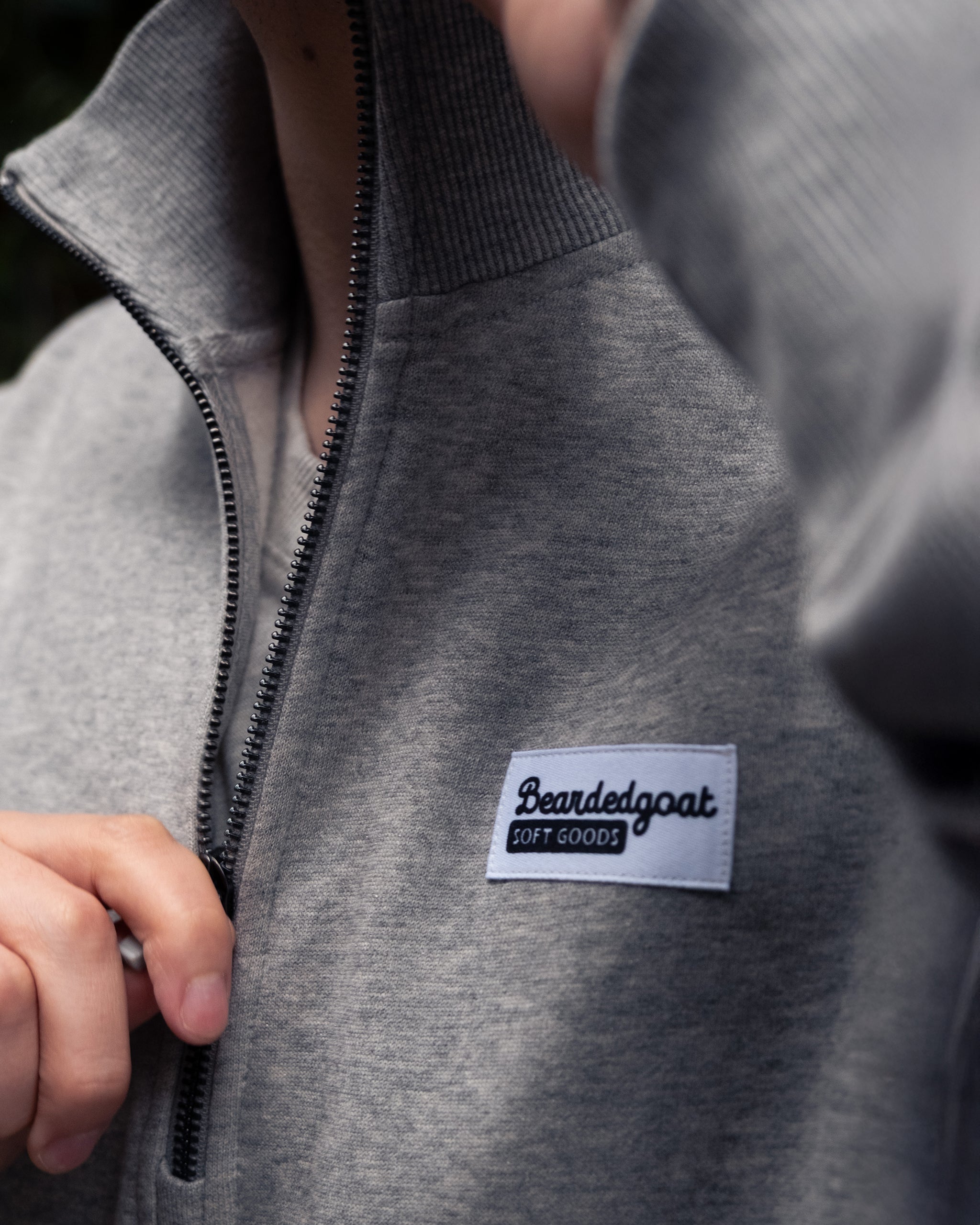 A person zips up the NEW — VOID™ Half Zip by BEARDEDGOAT, a light gray heavyweight fleece with a ribbed collar and a Beardedgoat SOFT GOODS patch. Only part of the person's face and hand are visible.