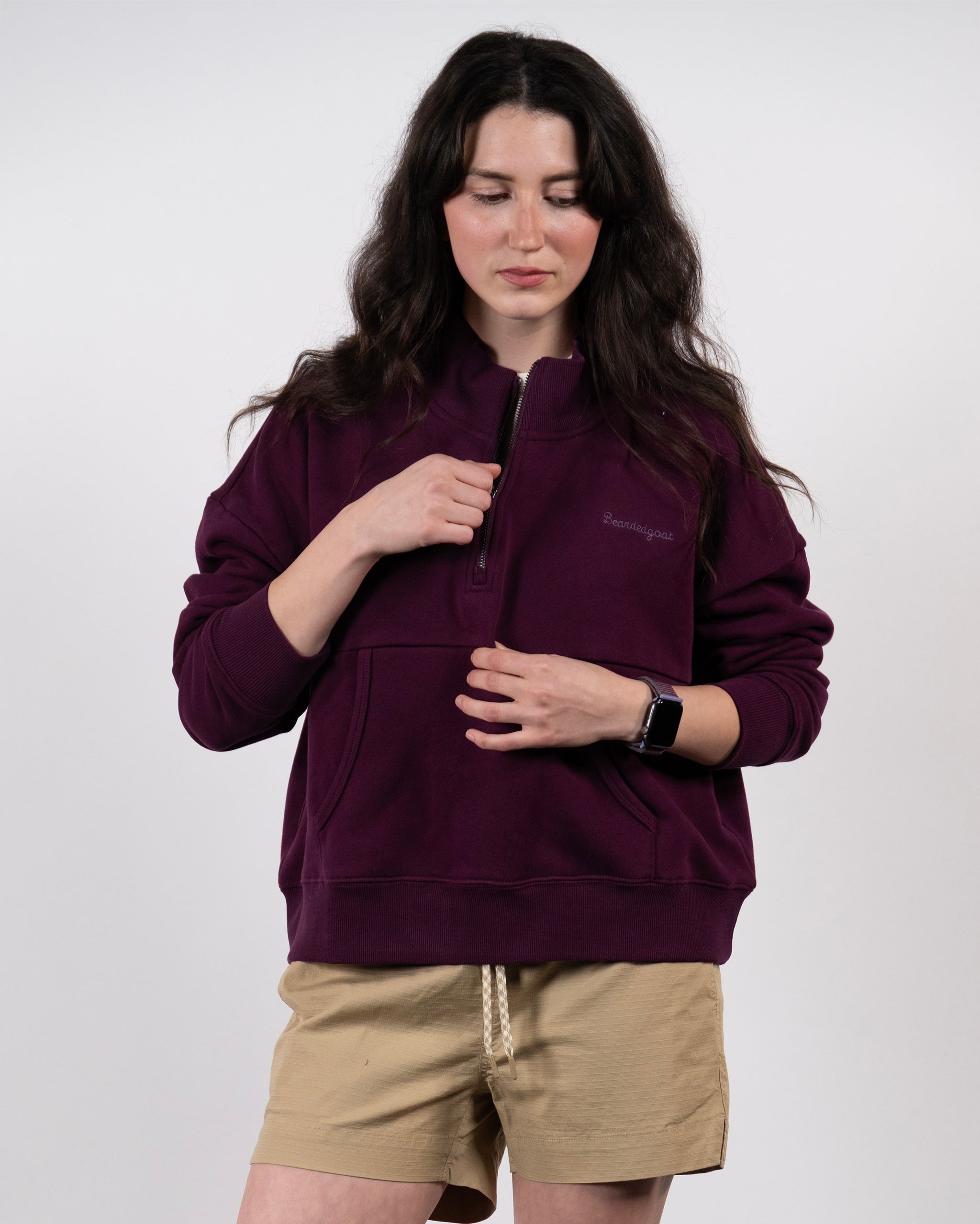 A person with long dark hair wears the BEARDEDGOAT NEW — Overnight™ Half Zip in maroon and khaki shorts, standing against a plain background while looking down and holding the zipper.