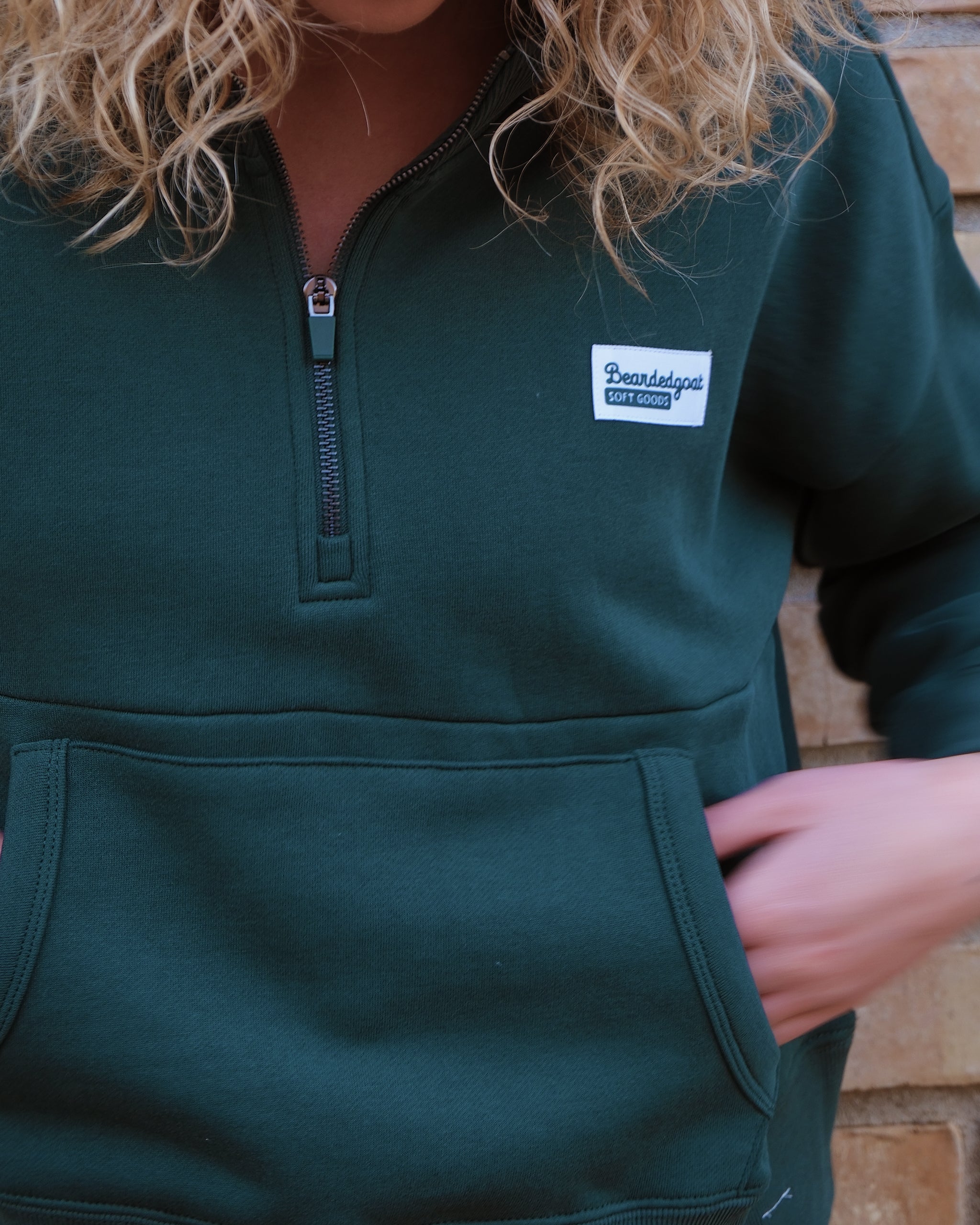 A person with curly blonde hair wears a dark green BEARDEDGOAT NEW — Overnight™ Half Zip fleece, featuring a Fort Collins patch, standing against a brick wall. Only the lower part of their face is visible.