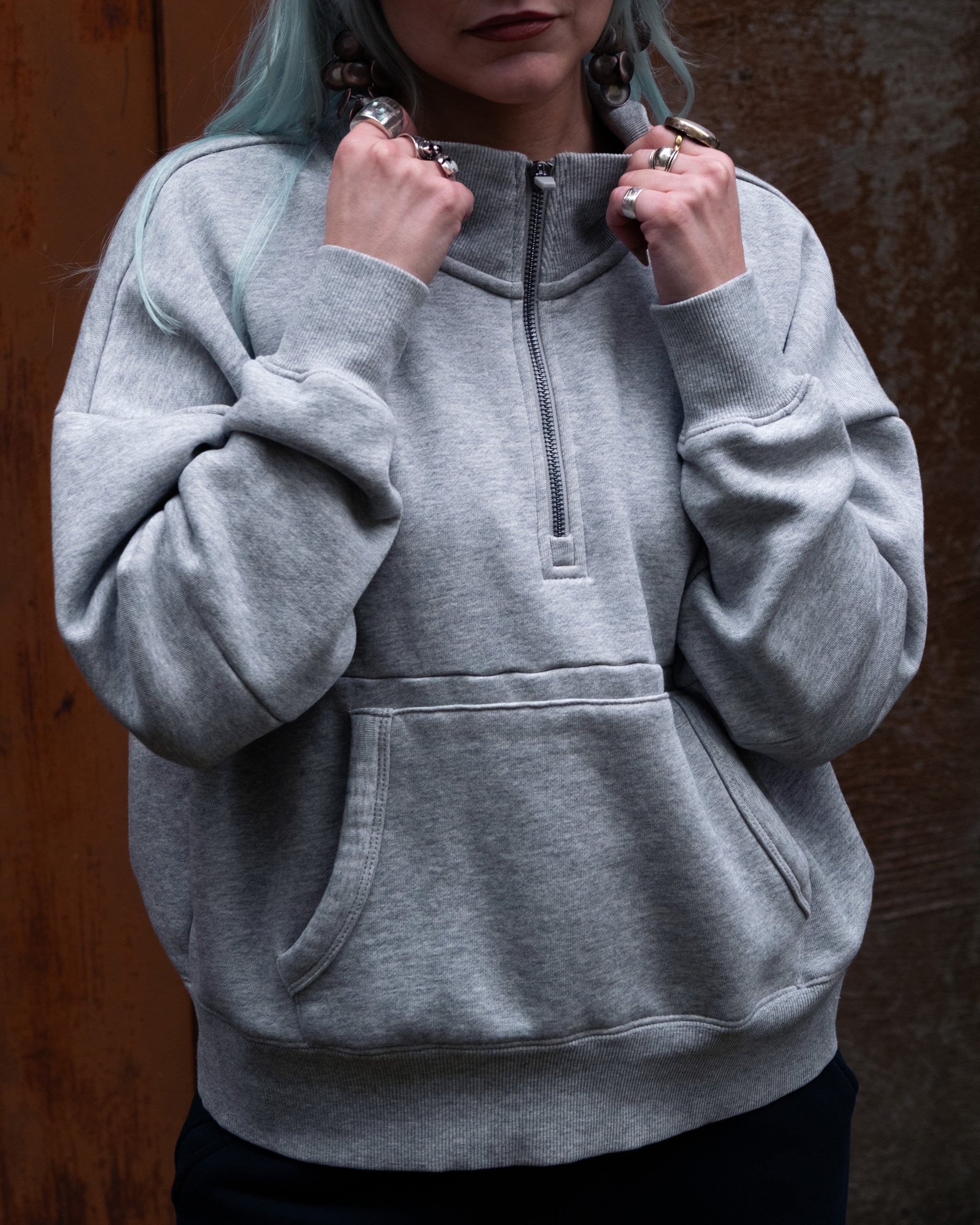 A person with long light blue hair wears the BEARDEDGOAT NEW — Overnight™ Half Zip, a loose-fitting gray premium cotton fleece sweatshirt, holding the collar to show off several rings. The backdrop is a brown textured wall.
