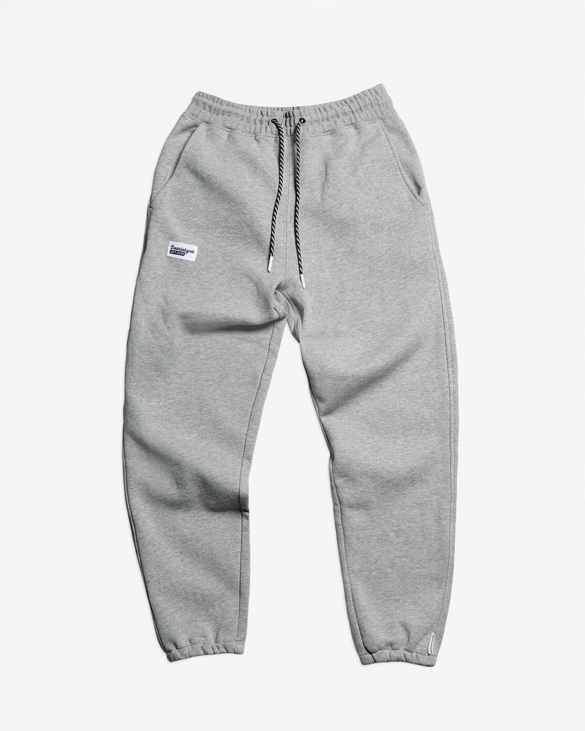 The NEW — Overnight™ Classic Jogger by BEARDEDGOAT features a light gray hue, high-rise waistband, black-and-white drawstring, elastic ankle cuffs, and a small white rectangular patch on the left pocket. Pants are shown laid flat on a white background.
