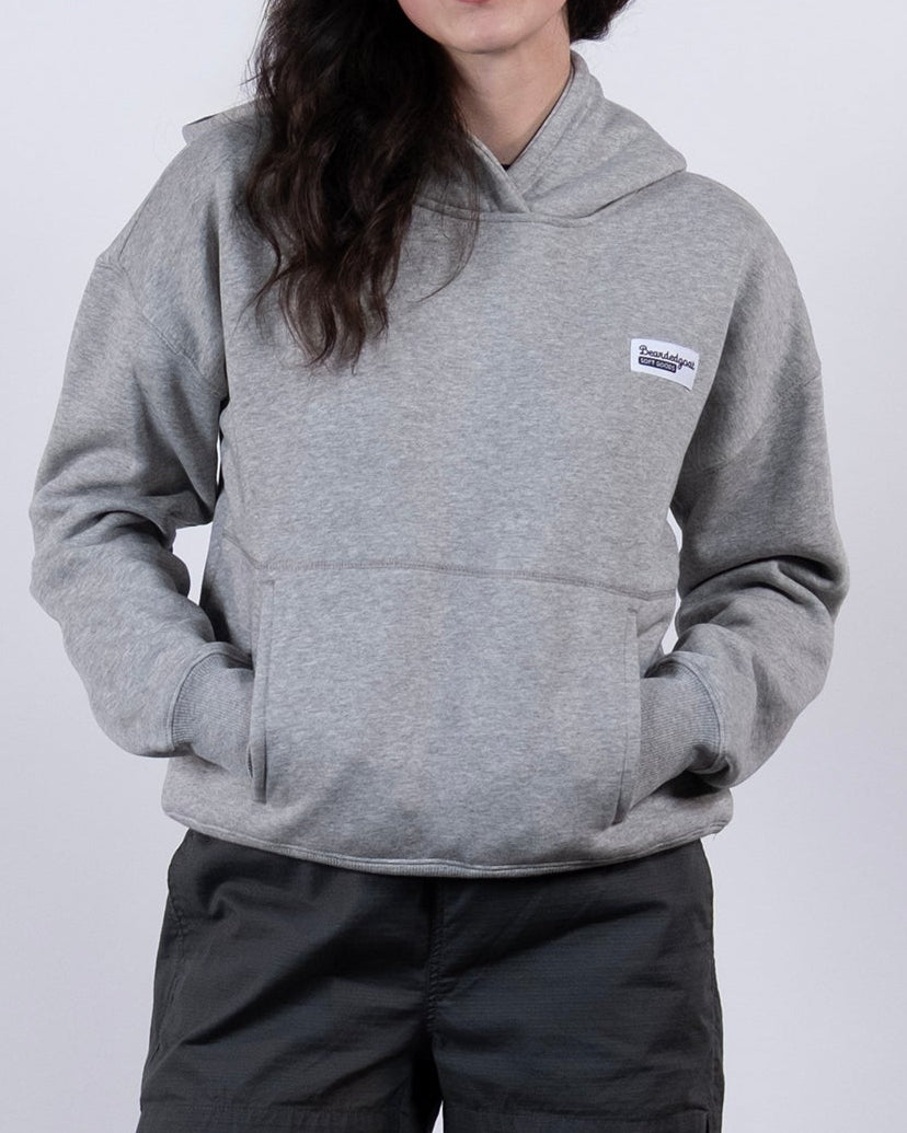 A woman with dark hair stands against a white background, wearing the beardedgoat Women's Overnight™ Hoodie in gray, paired with dark shorts, white socks, and gray sneakers. Her hands are tucked into the hoodie’s pockets.