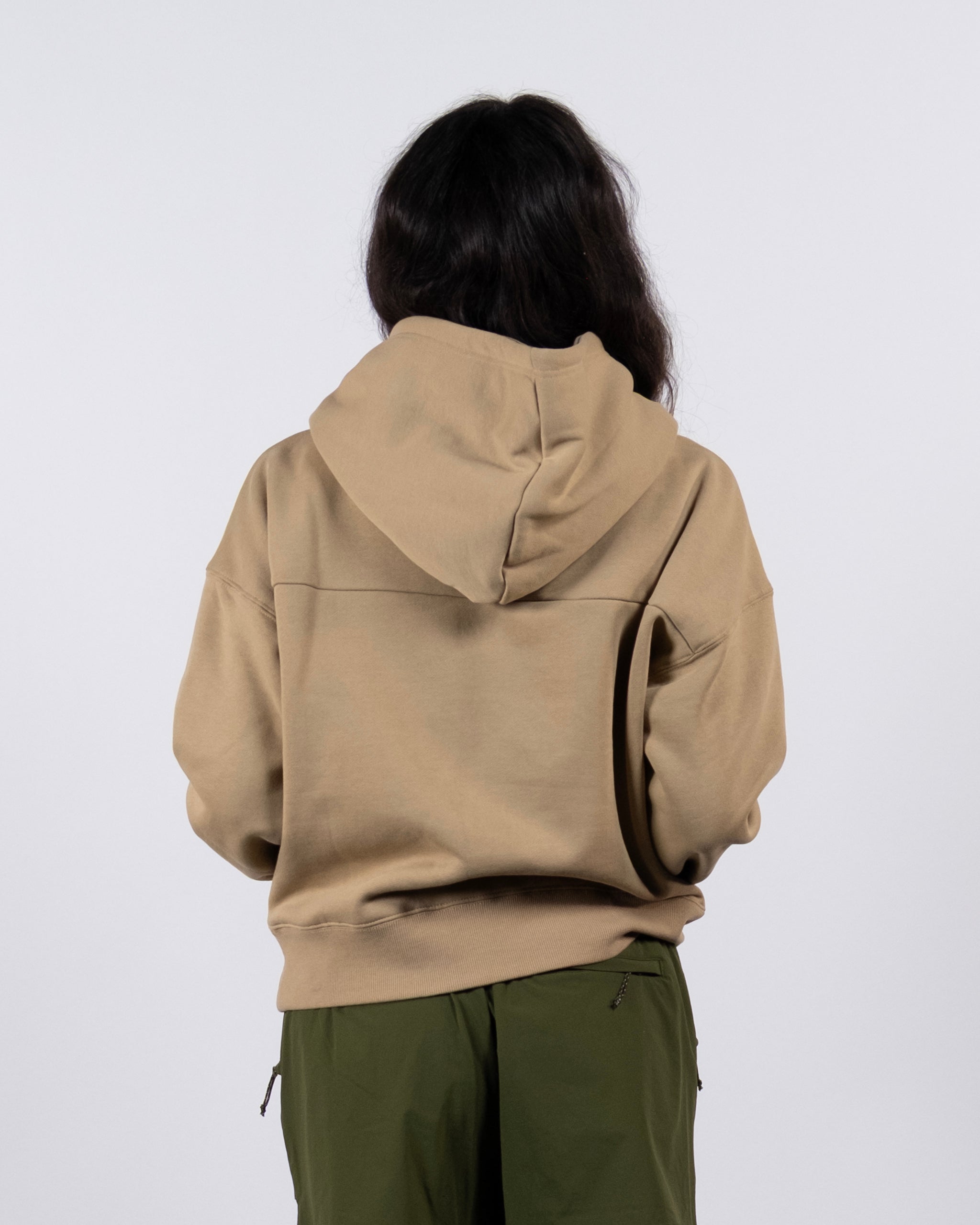 A person with long dark hair, seen from behind, wears a tan beardedgoat Women's Overnight™ Hoodie and olive green pants, standing against a plain light gray background.