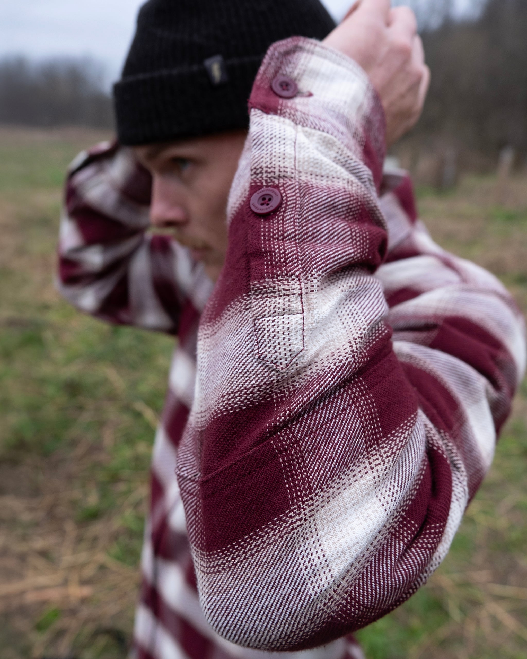 A person adjusts their hat outdoors in a grassy field while wearing the beardedgoat NEW--Ozark Utility Flannel™, highlighting the shirt's sleeve and button details.