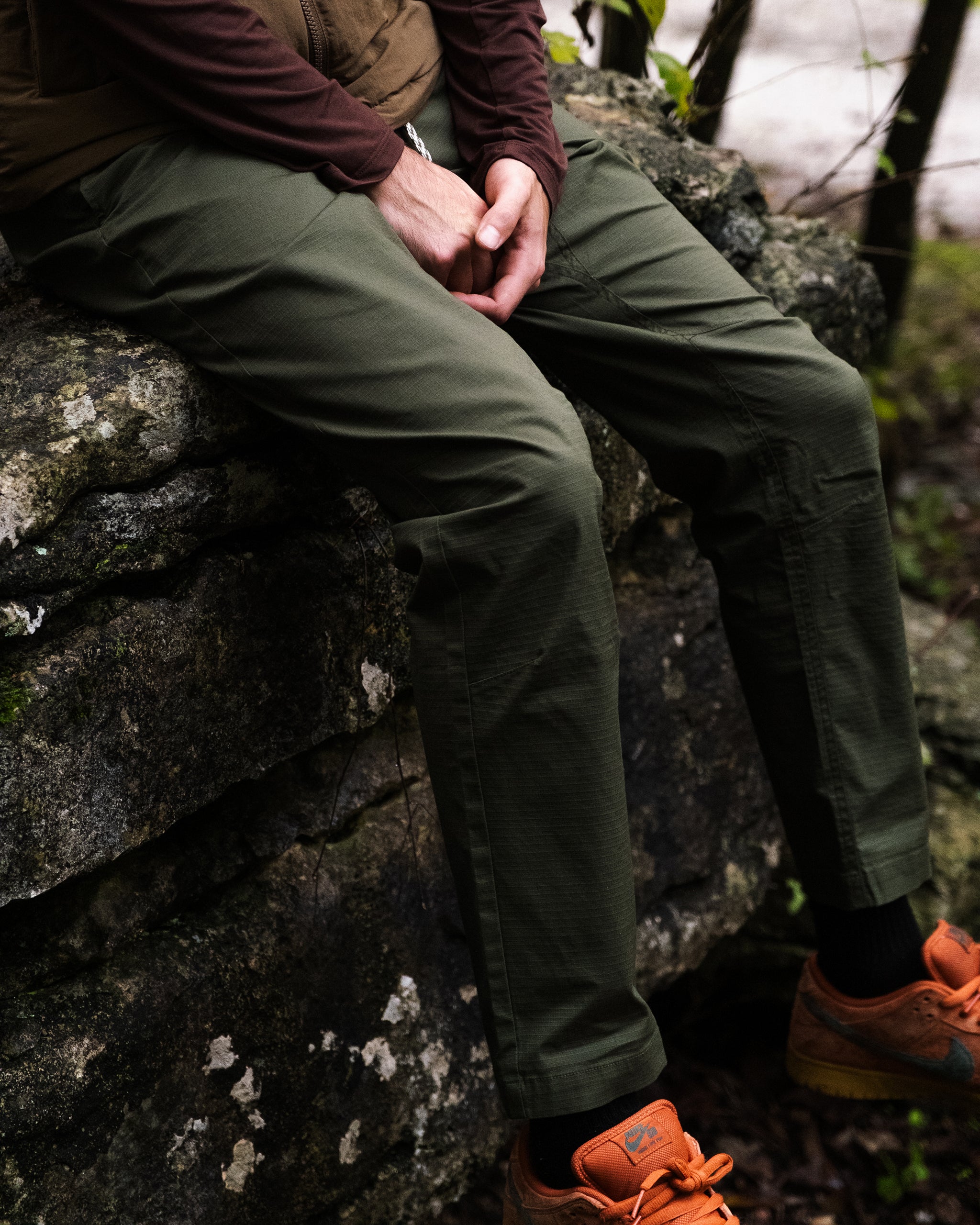 A person sits on a large rock outdoors, surrounded by greenery, wearing BEARDEDGOAT Men's Canopy Pant in olive green stretch cotton ripstop and orange shoes, with hands clasped.