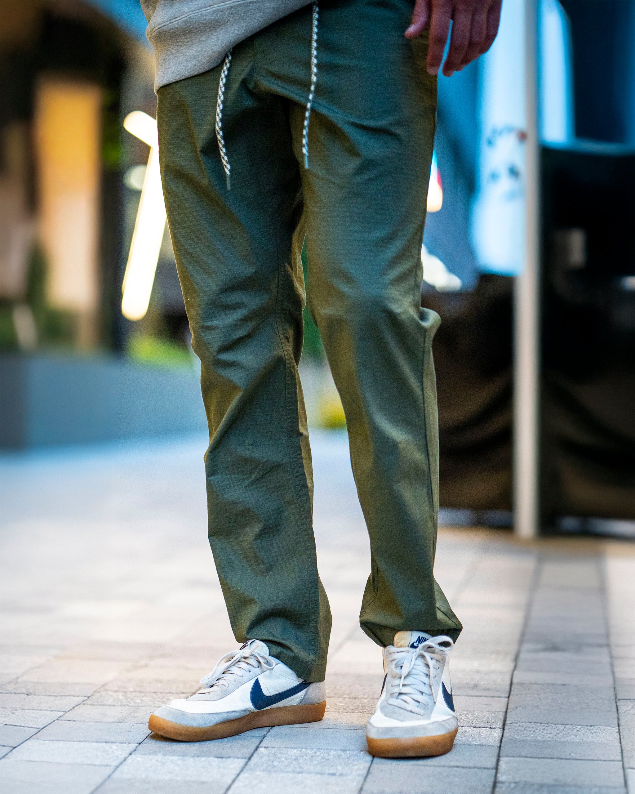 A person stands on a tiled walkway wearing BEARDEDGOAT Men's Canopy Pant in olive green, paired with white sneakers featuring navy swooshes and tan soles. Only a hand and part of a grey hoodie are visible; the upper body is mostly out of frame.