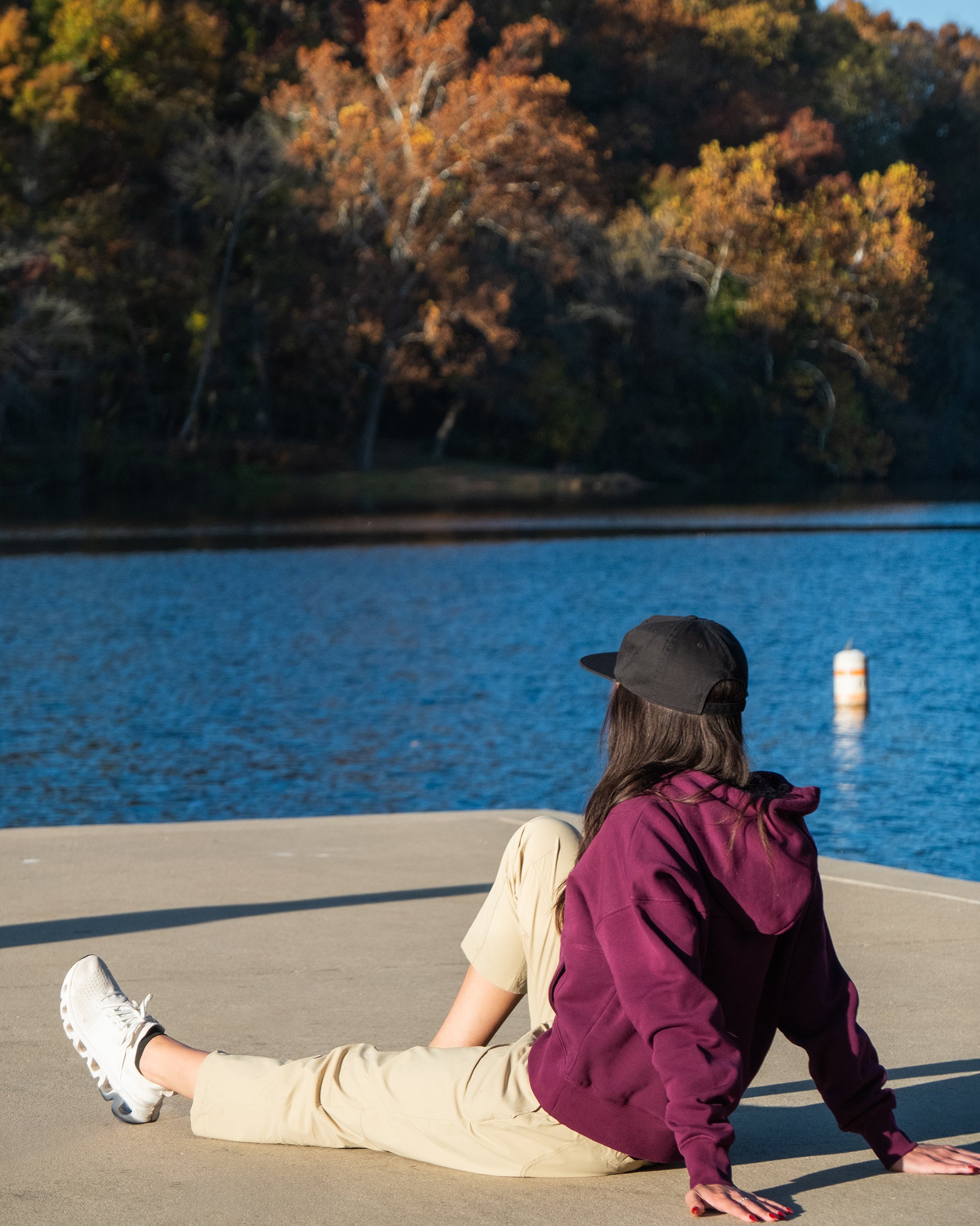 A person in BEARDEDGOAT’s stain-resistant, water-repellent Summit™ Pant, a burgundy hoodie, and a black cap sits on a dock by a lake, facing away. Colorful autumn trees are visible across the water behind them.