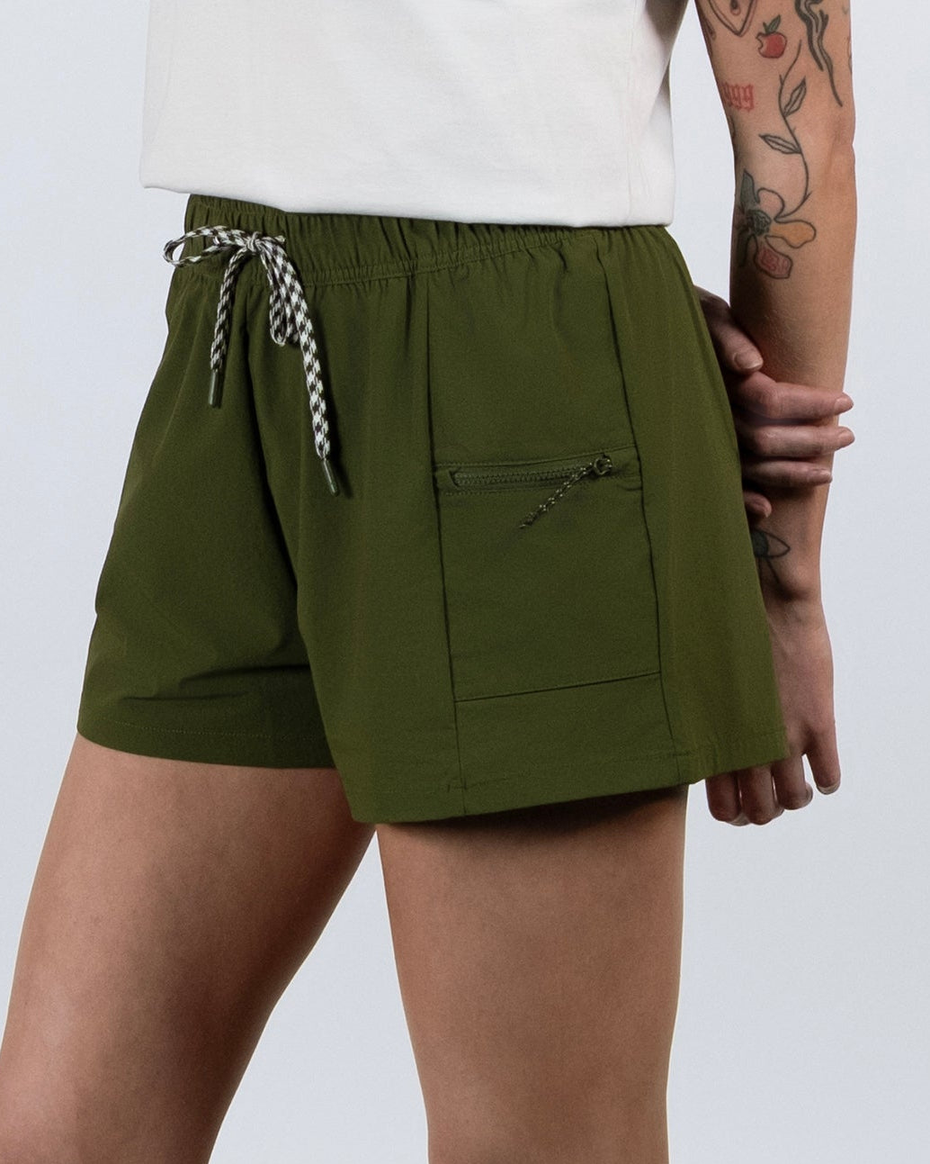 A person stands on a gray floor wearing BEARDEDGOAT Summit Shorts in olive green, crafted from lightweight ripstop with a front pocket and white drawstring. Only the lower half and one tattooed arm are visible, paired with white socks and sneakers.