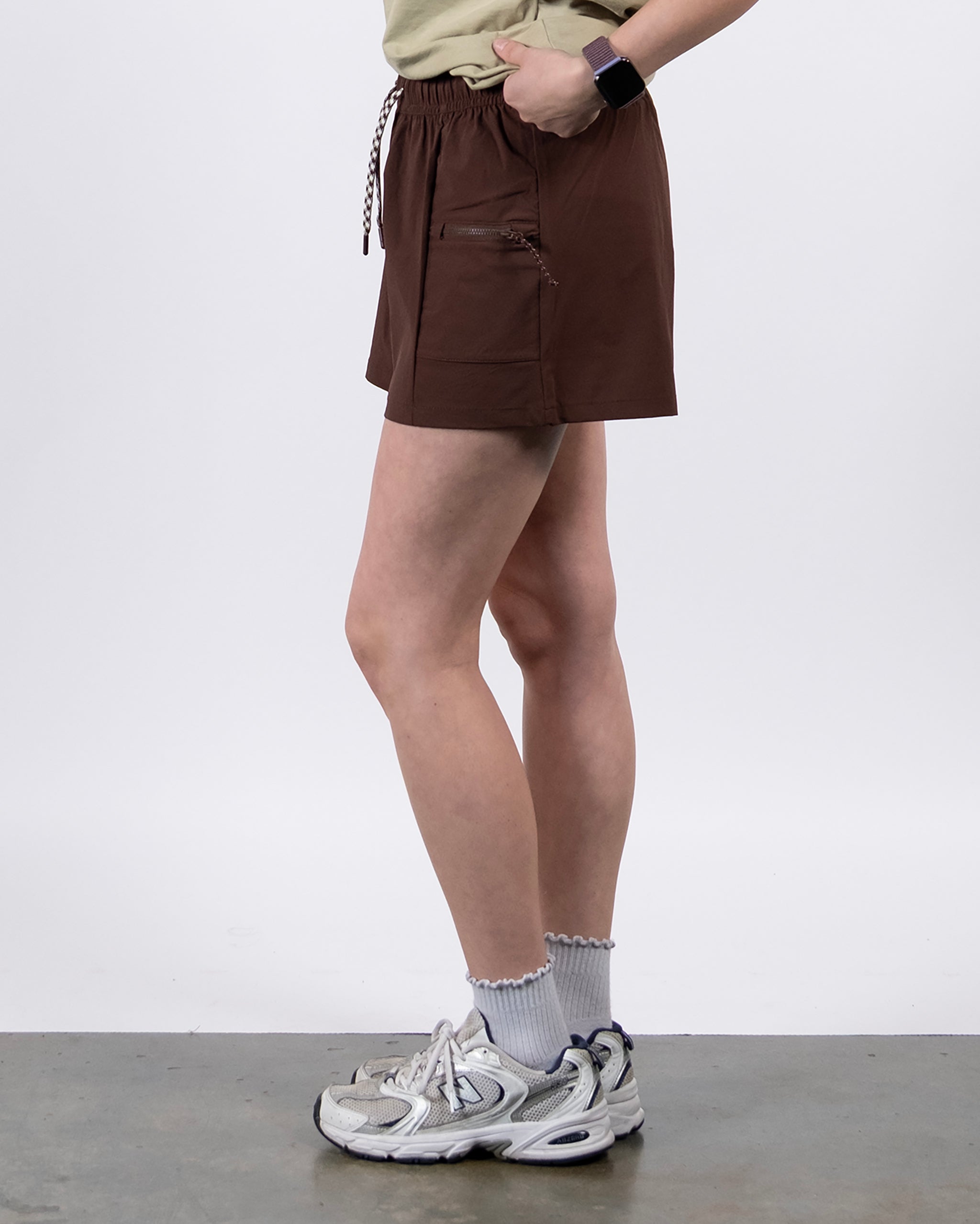 A person stands sideways against a white background wearing brown BEARDEDGOAT Summit Short trail shorts, a light beige t-shirt, frilly socks, white athletic shoes, and a black smartwatch. The shorts are made from lightweight ripstop fabric.