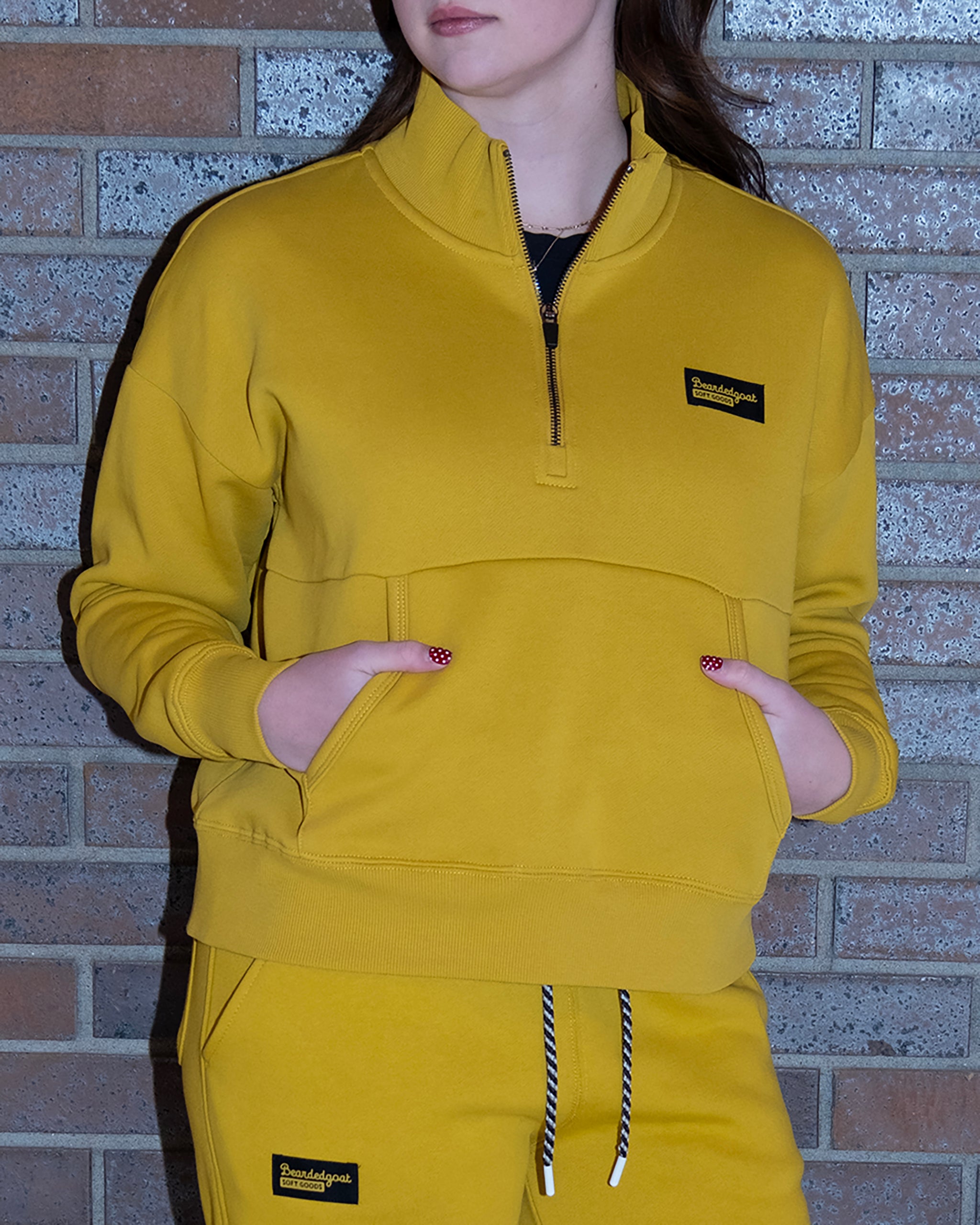 A person stands against a brick wall wearing the BEARDEDGOAT NEW — Overnight™ Half Zip sweatshirt and matching pants in yellow, made from premium cotton fleece, featuring black patches with yellow text and a black-and-white drawstring.