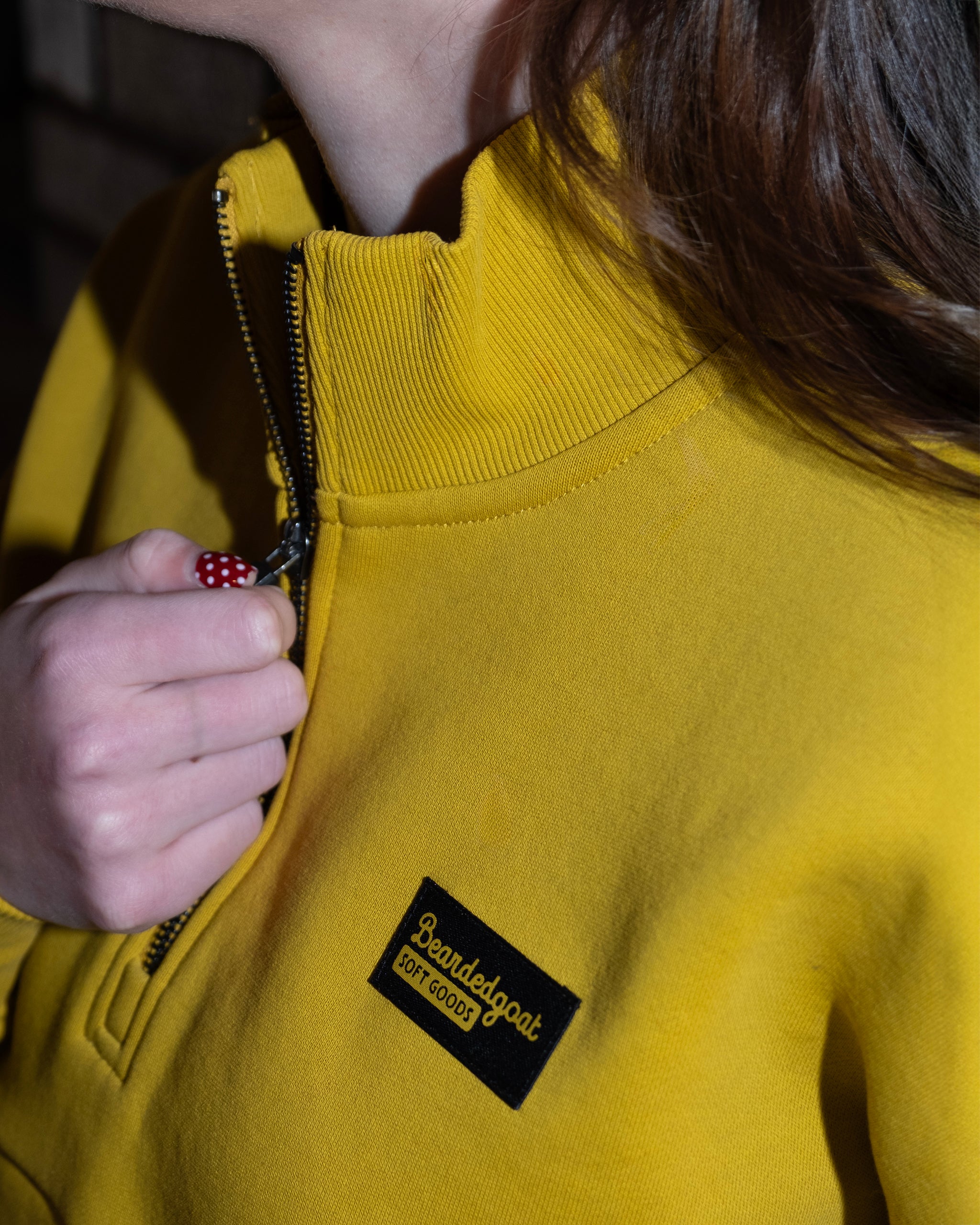 A person pulls up the zipper on a bright yellow BEARDEDGOAT NEW — Overnight™ Half Zip fleece. Only their lower face, neck, and hand are visible.