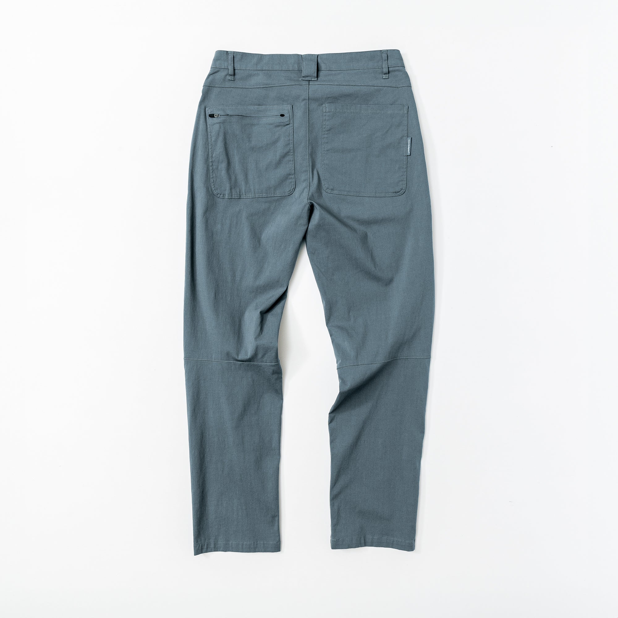 The Mens Union Pant by beardedgoat in gray-blue is shown from the back, laid flat on a white surface, highlighting two large utility pockets, a small tag on the right pocket, and knee seam details.