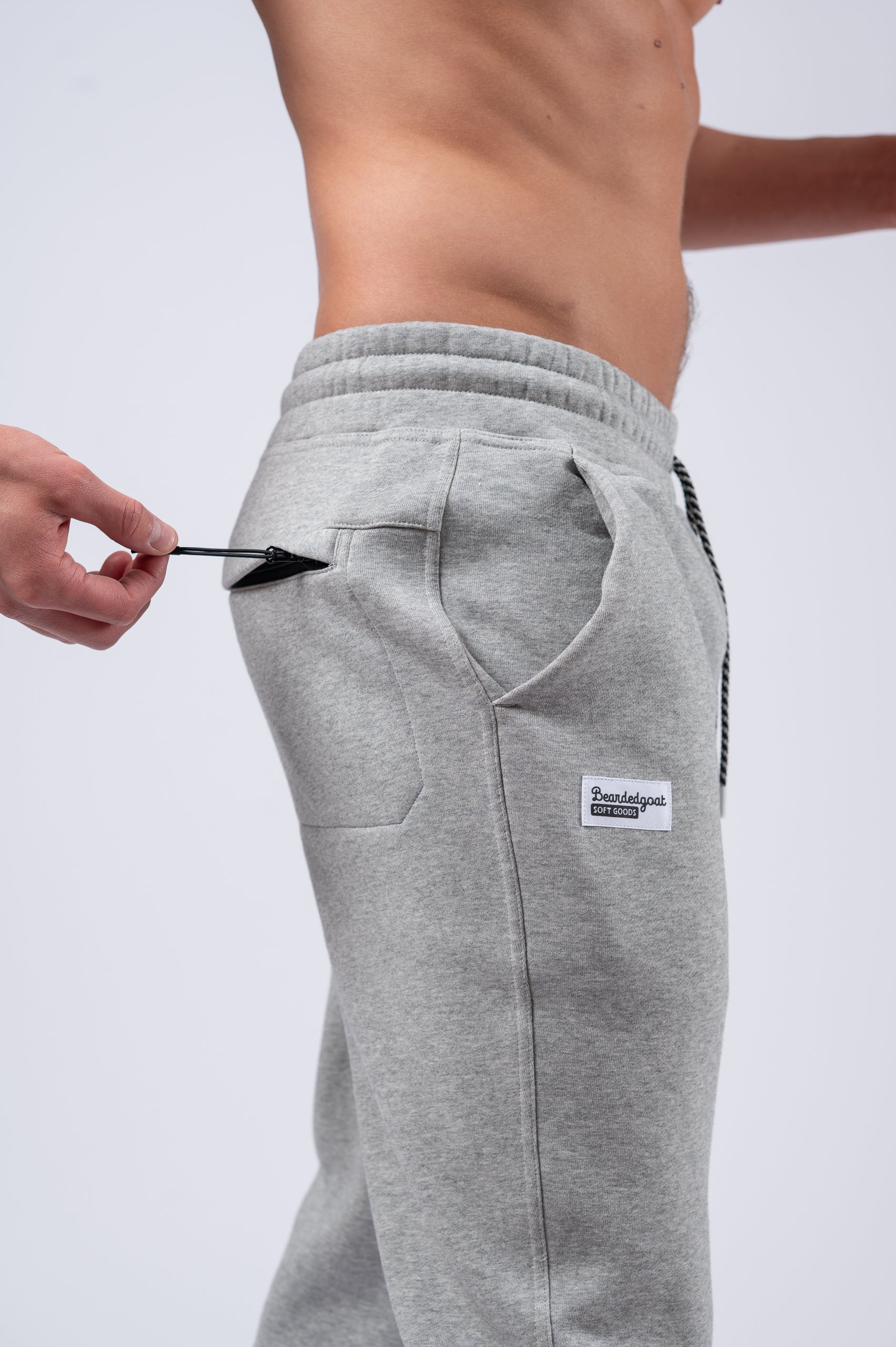 A person in beardedgoat Mens Void Sweatpants, gray, with a hand pulling the side pocket zipper; photo cropped to show just the lower torso and part of one arm.