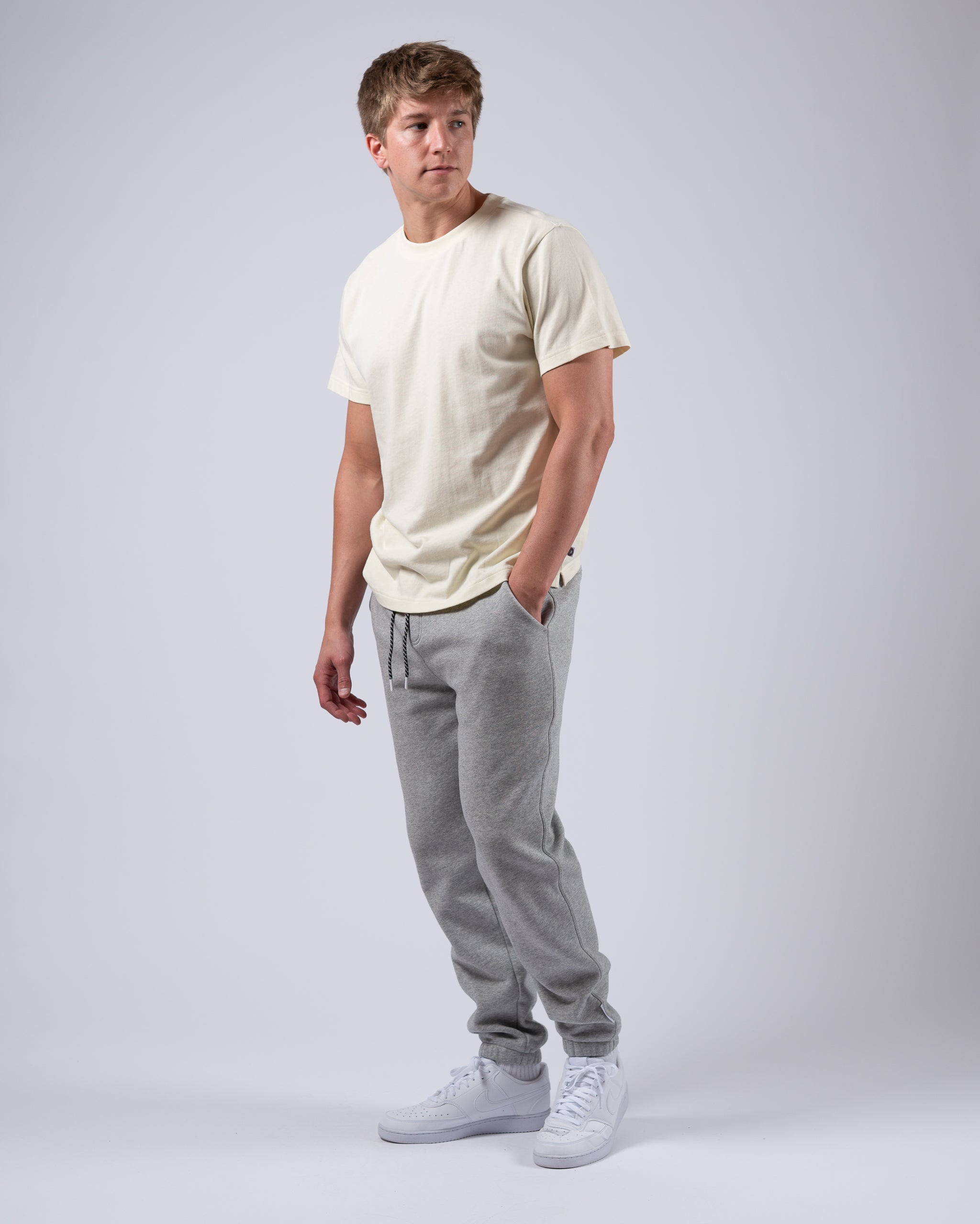 A young man stands against a plain light gray background, wearing a cream T-shirt, beardedgoat Mens Void Sweatpants with a drawstring, and white sneakers. He gazes slightly to the side with a neutral expression.