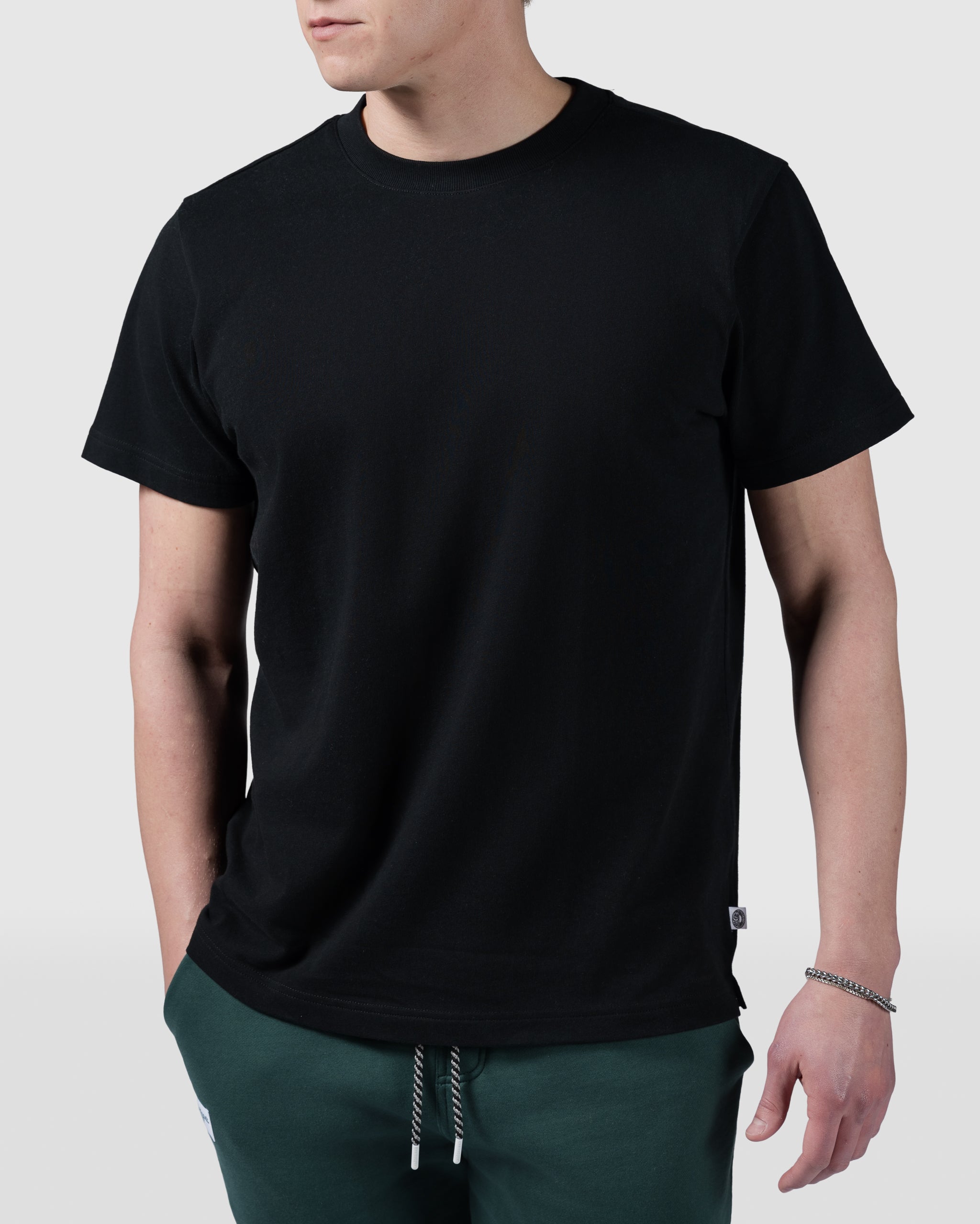 A person wearing the BEARDEDGOAT Mens Valley Tee in plain black and green pants stands with one hand in their pocket against a light gray background.