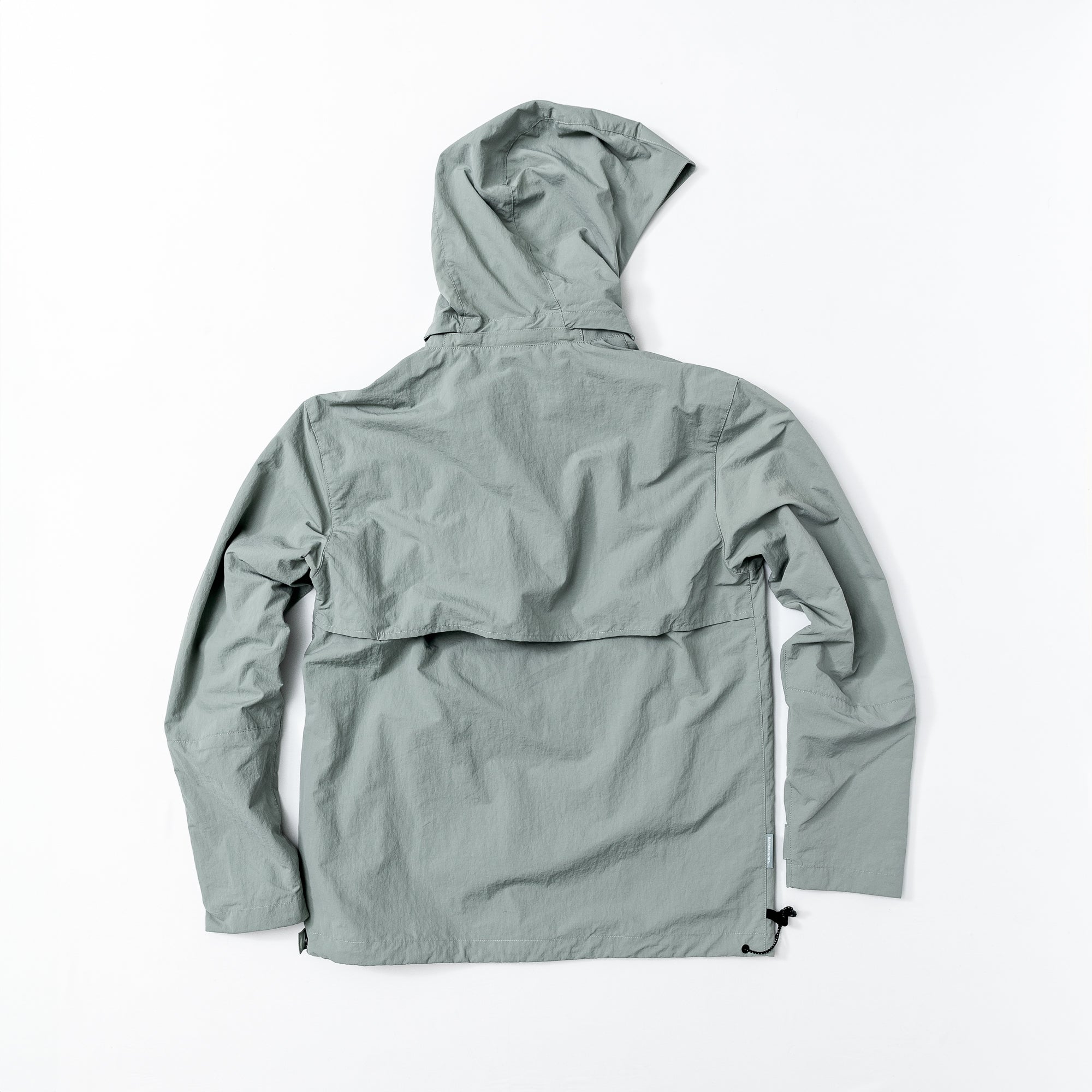 The beardedgoat Mens Deluge Packable Pullover, shown laid flat from the back, features a light gray-green, water-resistant fabric with long sleeves, loose fit, upper back vented flap, and a lightweight, slightly crinkled texture.