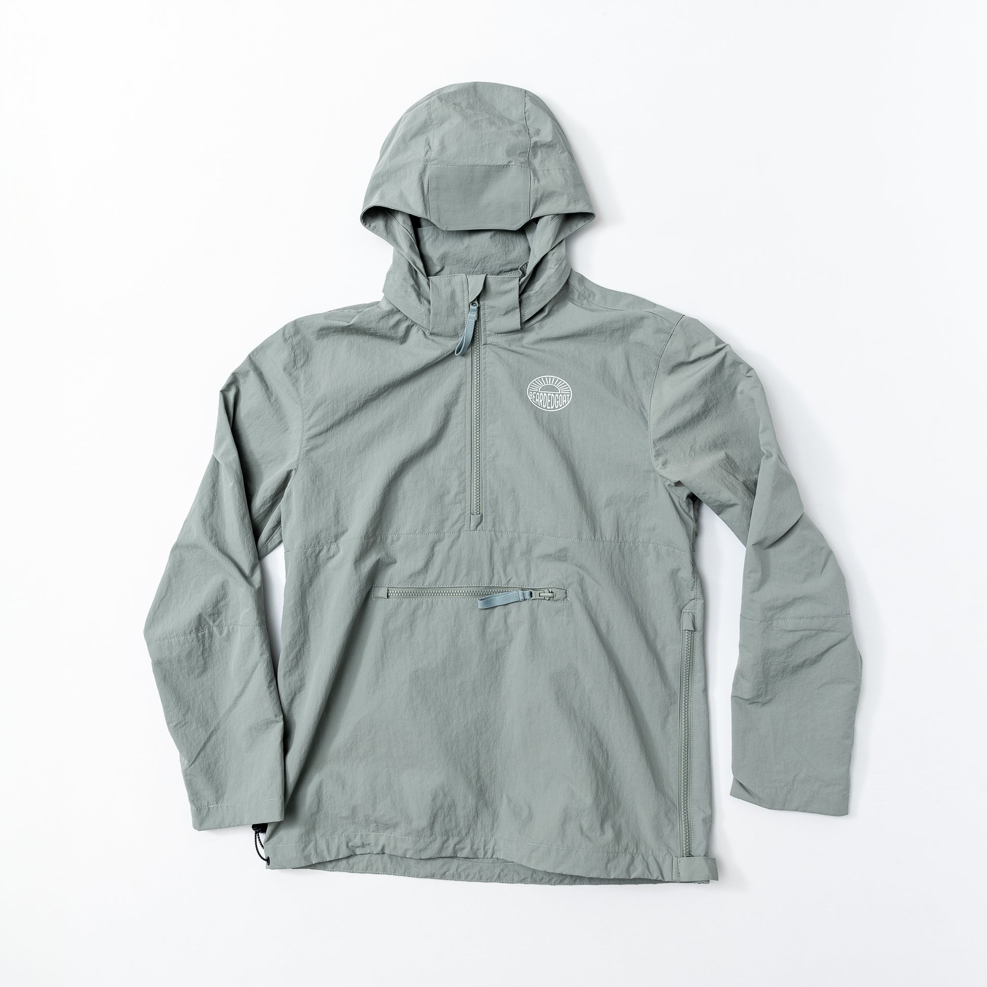 The beardedgoat Mens Deluge Packable Pullover is a light gray, water-resistant windbreaker with a half-zip front, zippered chest pocket, and circular logo—an adventure-ready layer shown flat on white.