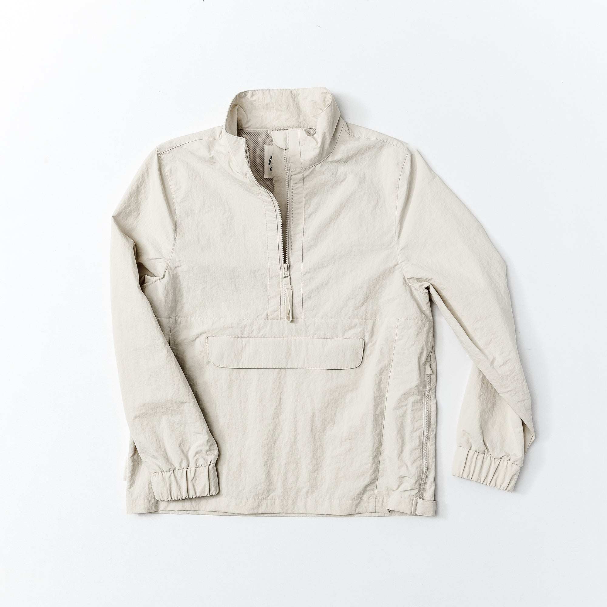 The beardedgoat Womens Sunchase Packable Pullover is a light beige, long-sleeve half-zip jacket with a front flap pocket, elastic cuffs, and a standing collar, shown flat on a white background.