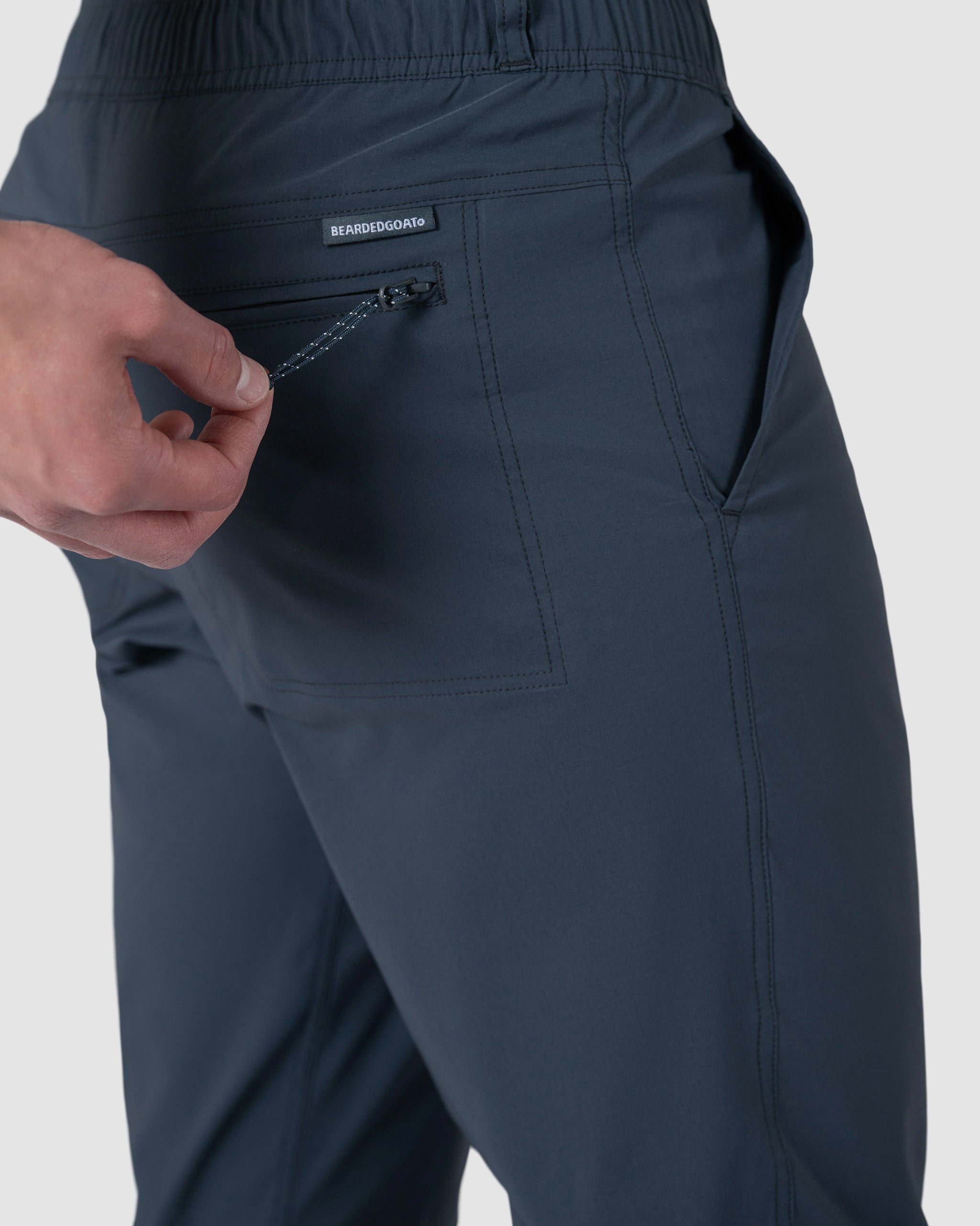 A man unzips a small back pocket on BEARDEDGOAT Mens Rover Pant in dark gray, featuring an elastic waistband and a label above the pocket against a plain light gray background.