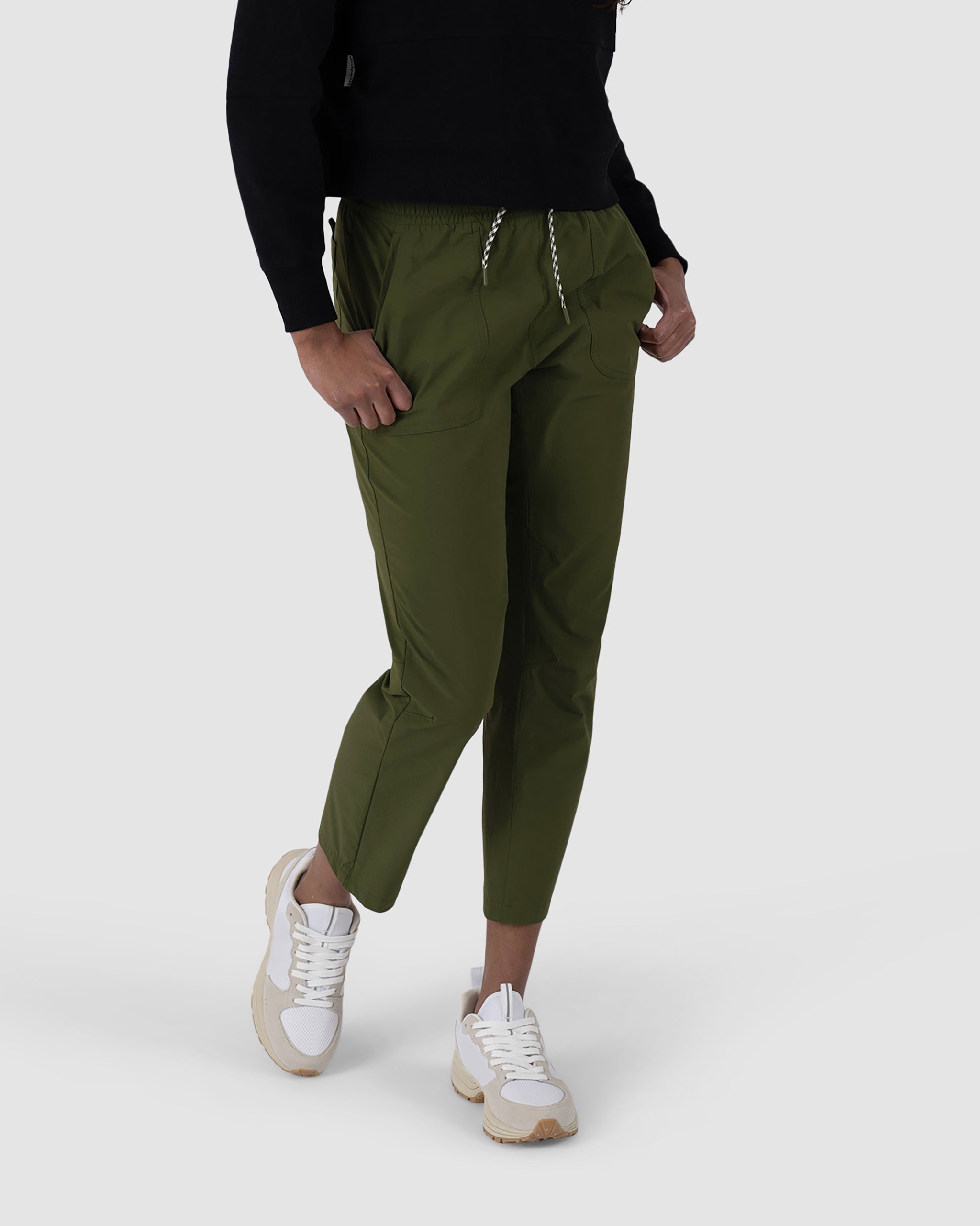 A person wearing BEARDEDGOAT NEW — Summit™ Pant in Deep Moss, a black top, and white sneakers with beige soles stands against a plain light gray background.