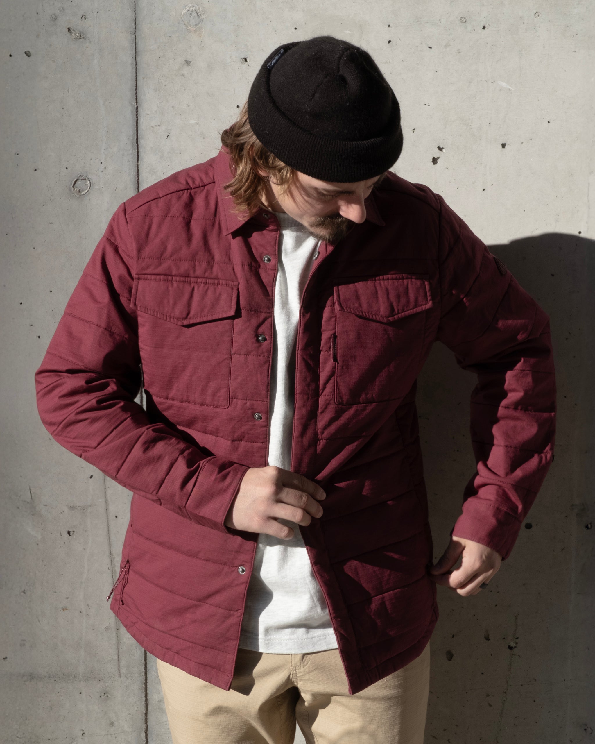 Someone wearing a maroon BEARDEDGOAT NEW — CANOPY™ Shacket, white shirt, beige pants, and black beanie stands against a concrete wall, looking down while adjusting the rugged shacket.