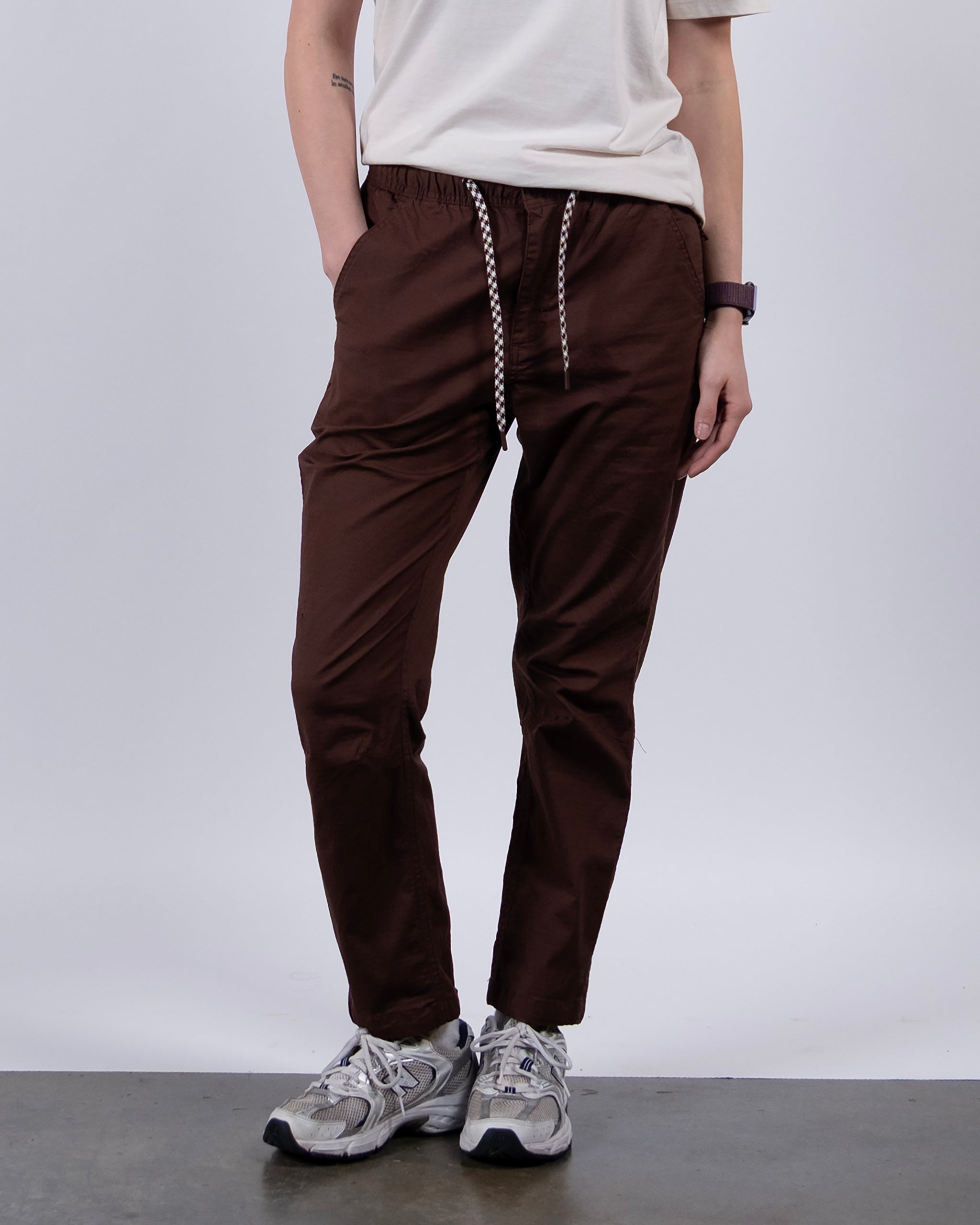 A person stands against a plain white background with hands in their pockets, wearing a white short-sleeve t-shirt, BEARDEDGOAT Women's Canopy™ Pant in stretch cotton ripstop with adjustable ankle drawcords, and gray sneakers.