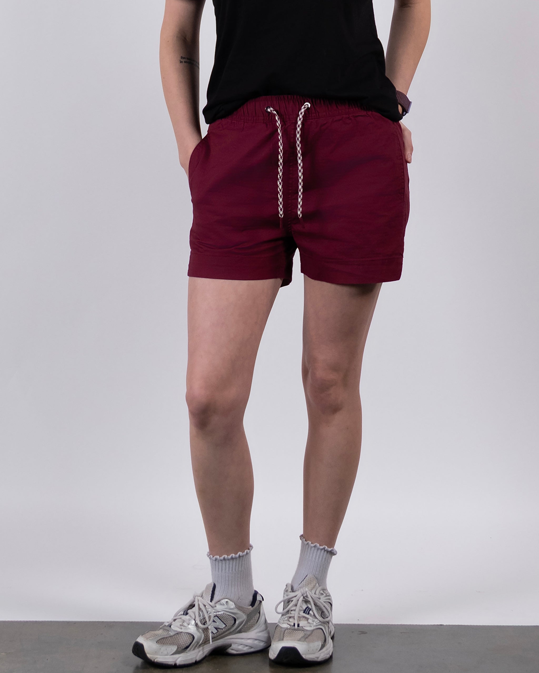 A person stands against a plain white background with hands in pockets, wearing a black t-shirt, BEARDEDGOAT Women's Canopy Shorts in maroon stretch cotton ripstop for breathable comfort, white ruffled socks, and gray athletic sneakers.