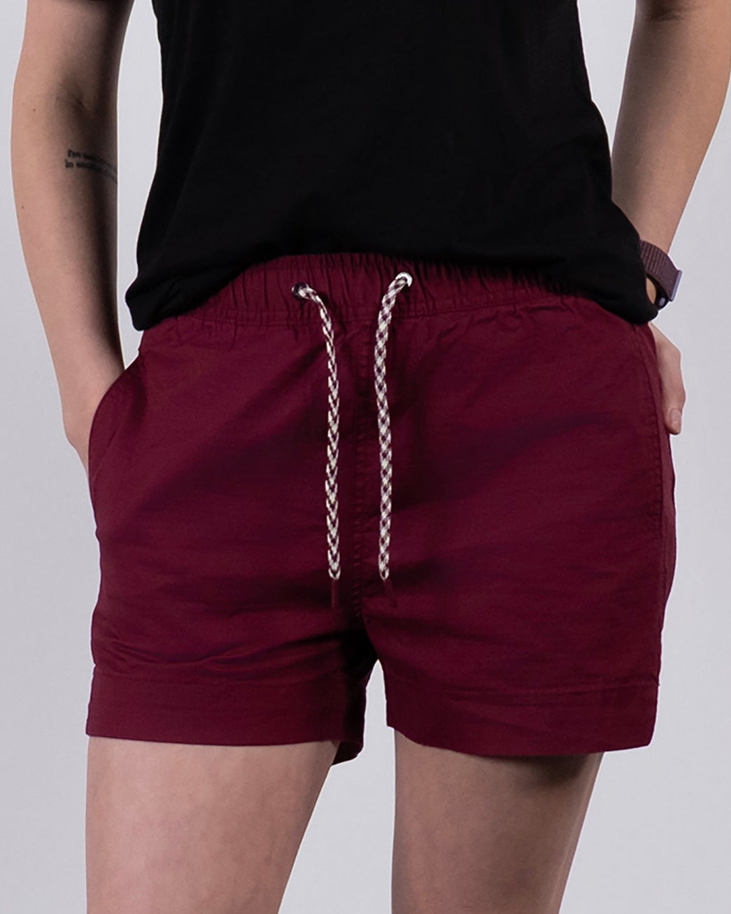 A person stands against a plain white background with hands in pockets, wearing a black t-shirt, BEARDEDGOAT Women's Canopy Shorts in maroon stretch cotton ripstop for breathable comfort, white ruffled socks, and gray athletic sneakers.
