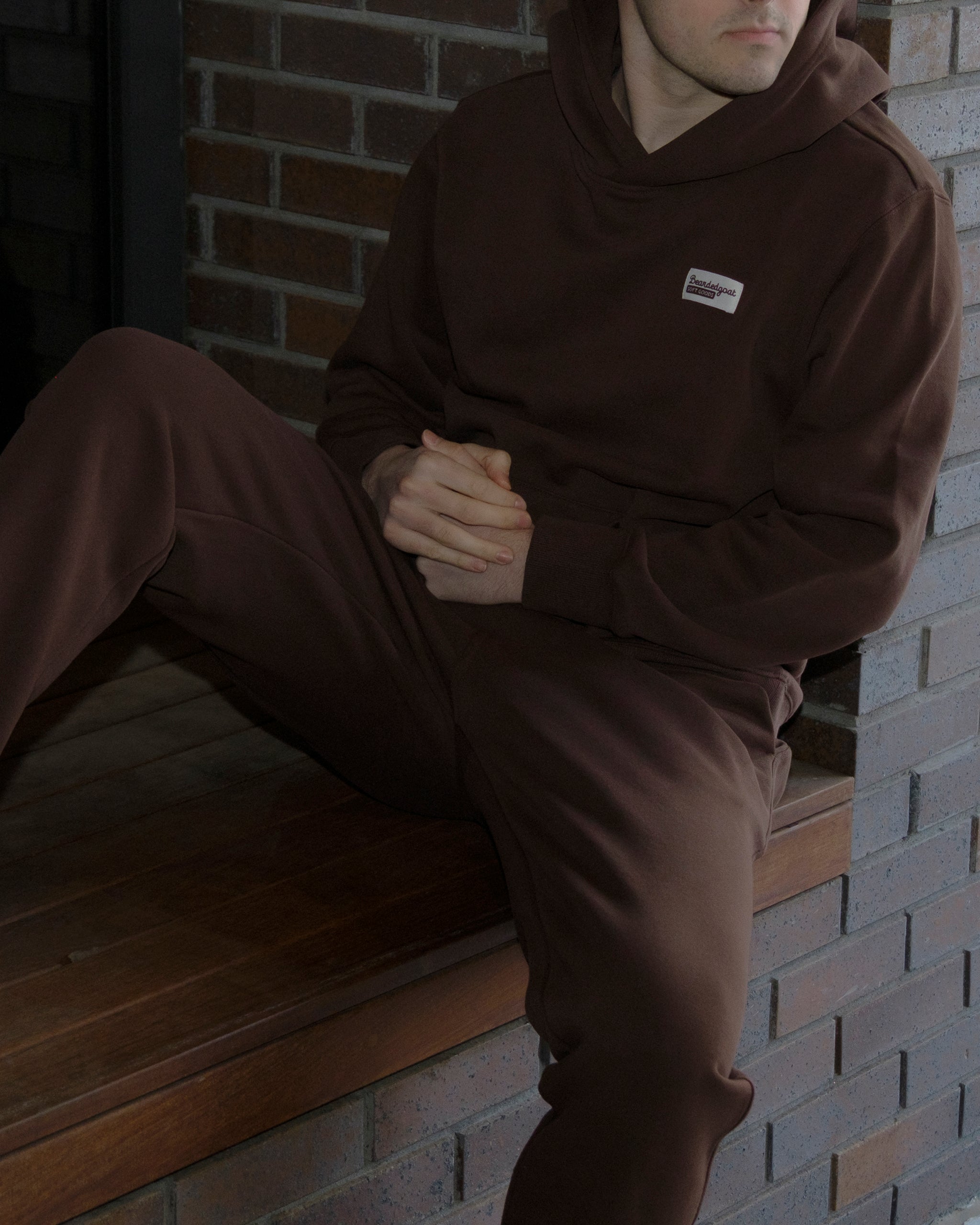 A person in the beardedgoat The VOID™ Hoodie and matching pants in heavyweight cotton fleece sits on a wooden ledge by a brick wall, hands clasped, face partly out of frame.