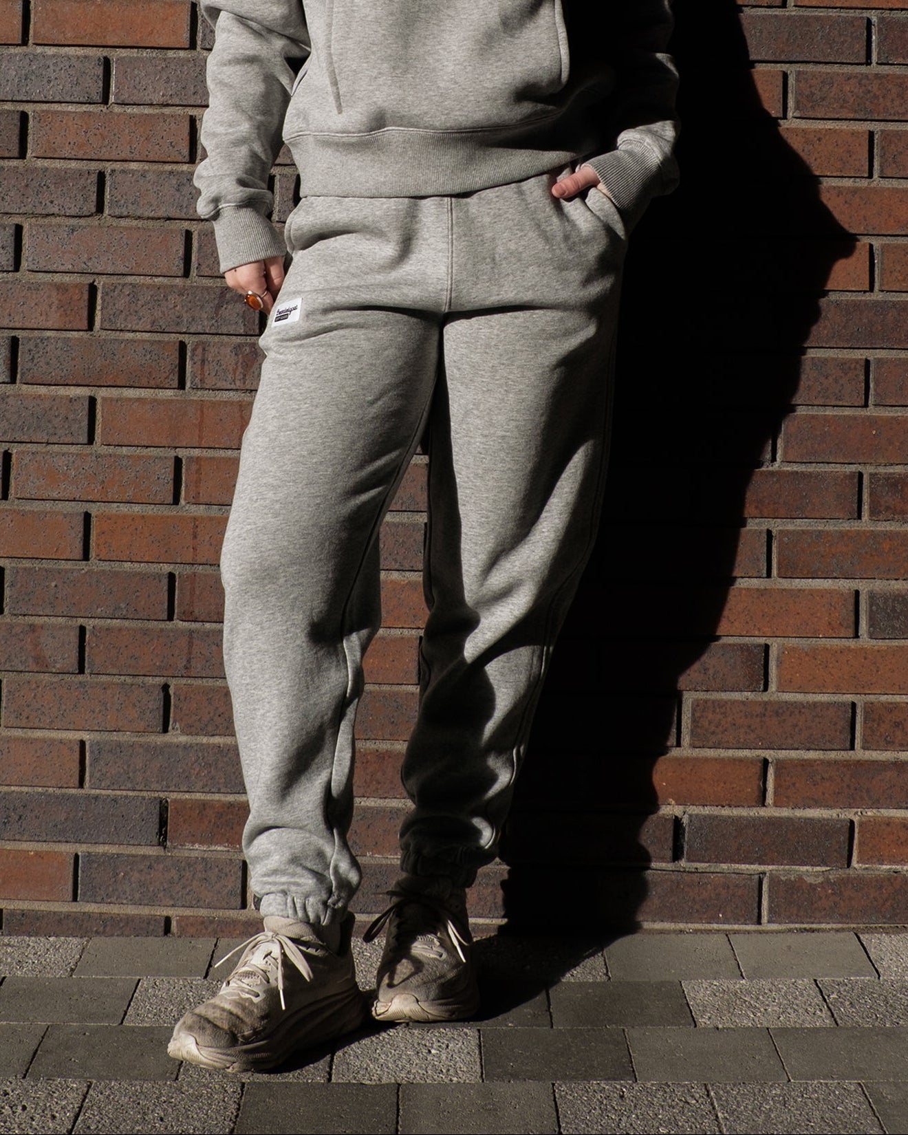 Wearing the BEARDEDGOAT NEW — Overnight™ Classic Jogger in light gray with a high-rise waistband and beige sneakers, a person stands against a brick wall with hands in pockets; their face is hidden, and shadows fall across the scene.