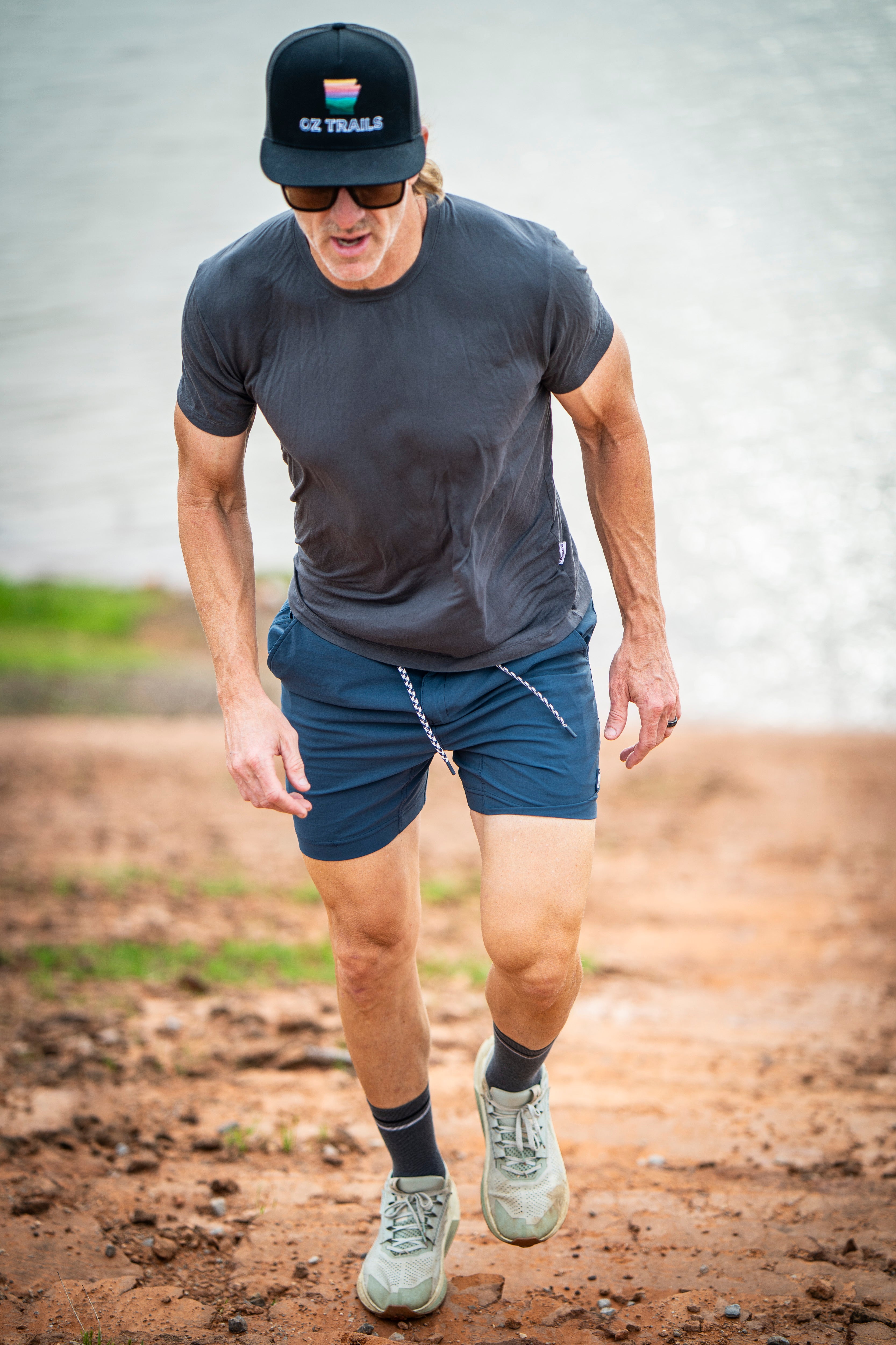 A man wearing the BEARDEDGOAT Men’s EverZen™ Short Sleeve, sunglasses, blue shorts, and sneakers hikes uphill on a sunny dirt trail near water, enjoying performance comfort in his bamboo blend shirt.