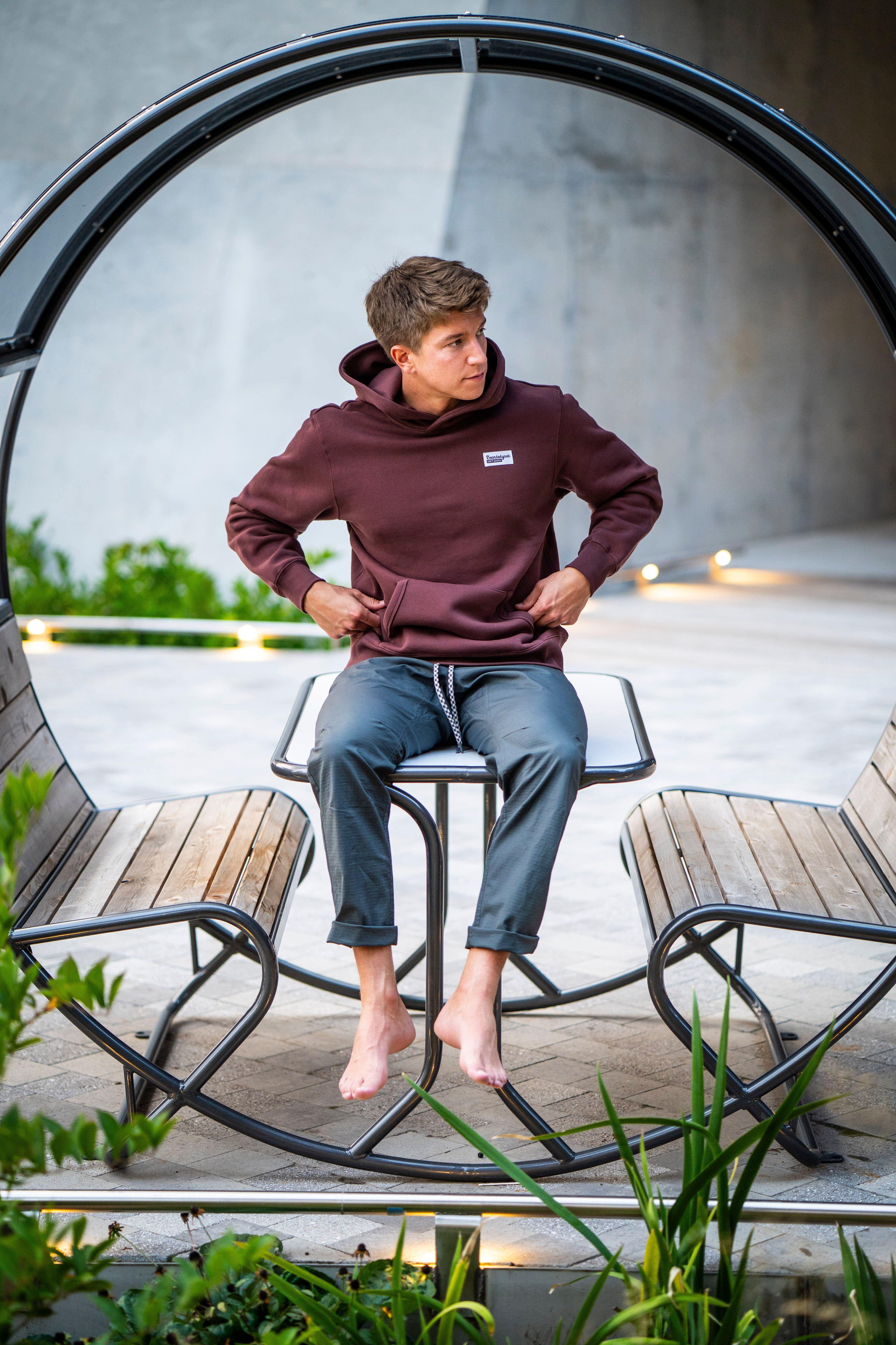 A barefoot young man in a maroon beardedgoat Men's Void Hoodie and gray pants sits on a modern circular bench. He looks to his left, with green plants in the foreground and soft lights illuminating the background.