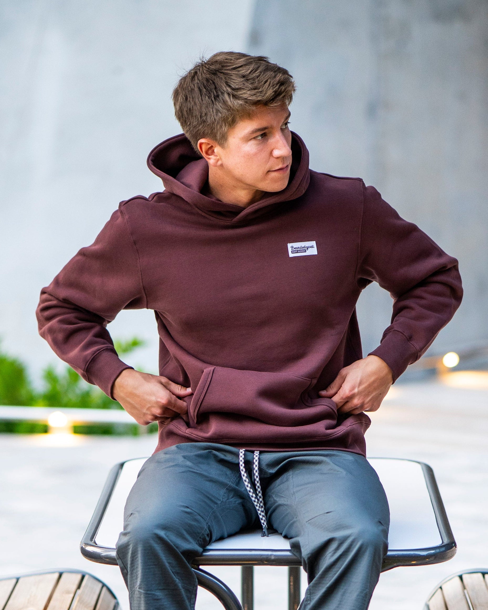 A barefoot young man in a maroon beardedgoat Men's Void Hoodie and gray pants sits on a modern circular bench. He looks to his left, with green plants in the foreground and soft lights illuminating the background.