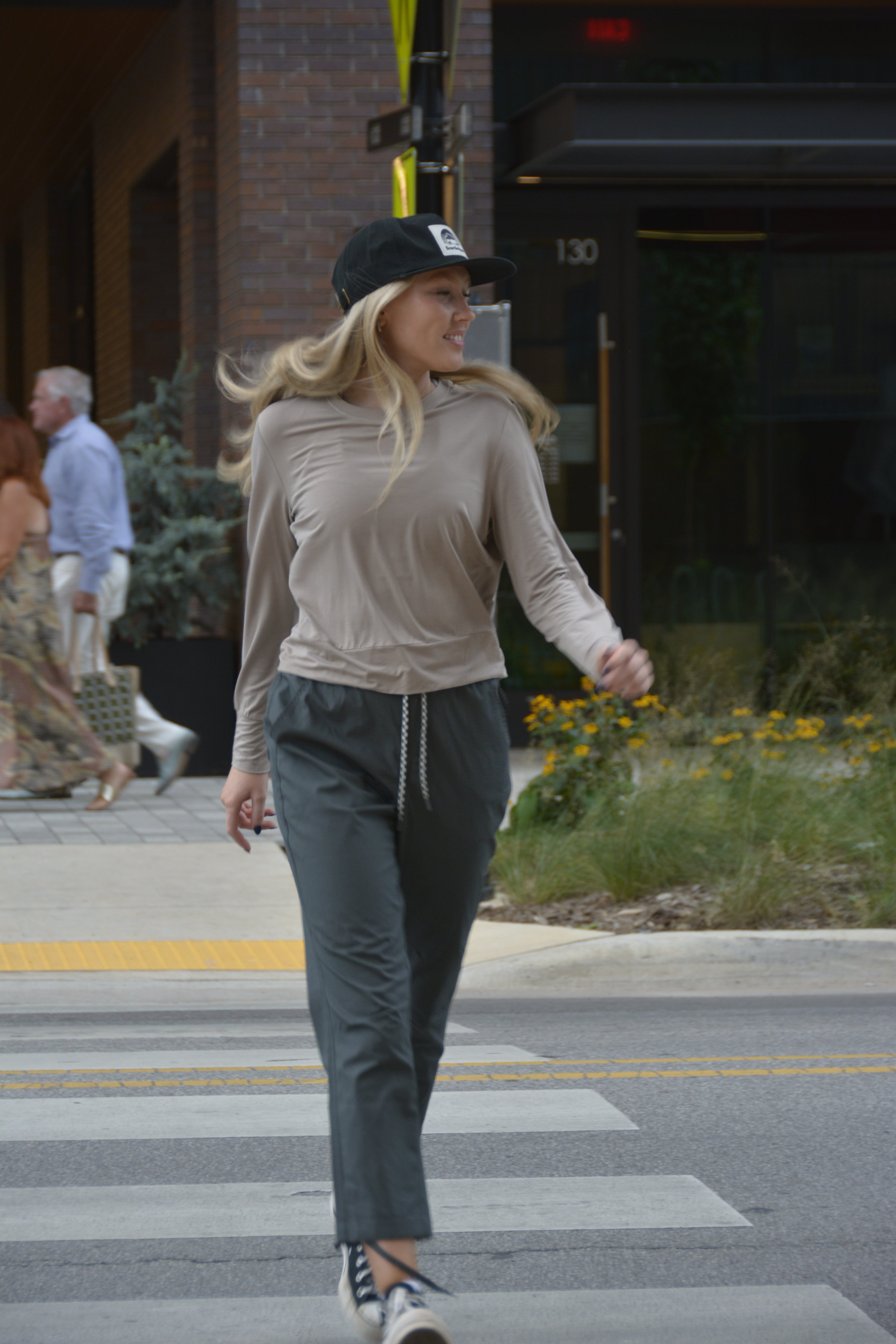 A woman in a BEARDEDGOAT Women’s EverZen™ Bamboo Long Sleeve, dark joggers, sneakers, and a black cap confidently crosses a city street, smiling and glancing left.