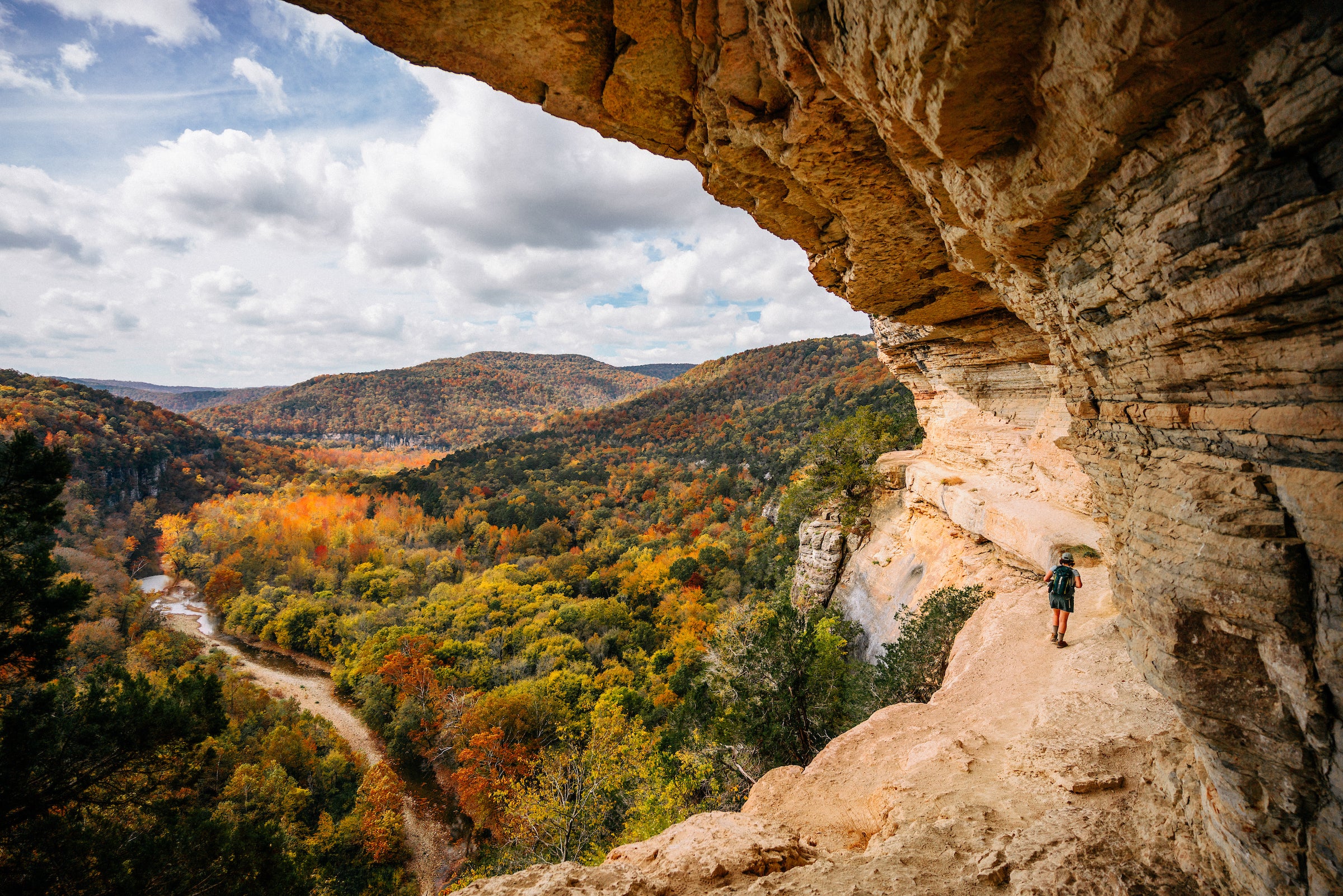 A hiker walks along a rocky cliff edge overlooking a vast, colorful forest valley with autumn foliage and distant hills under a partly cloudy sky.