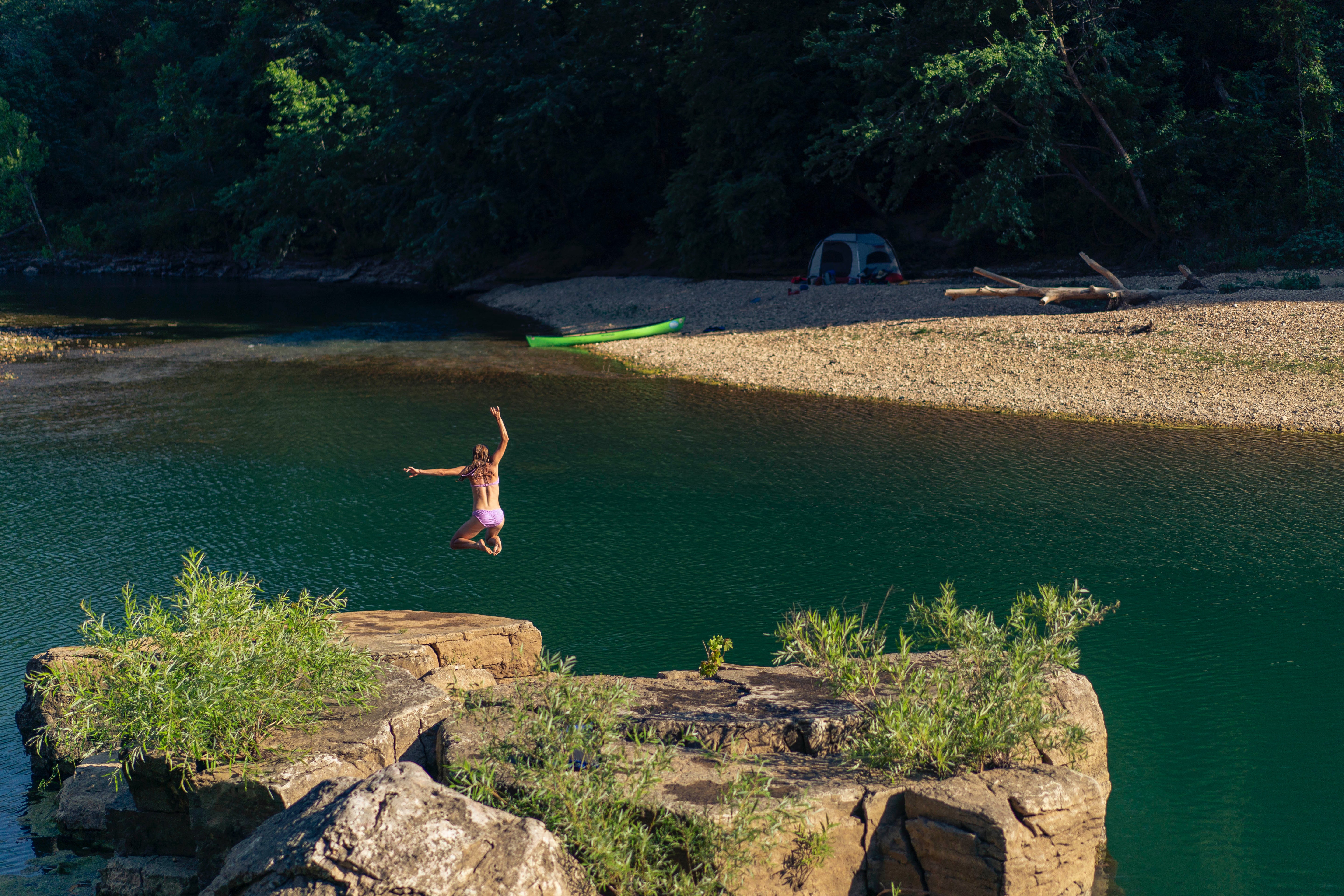 A person in swimwear jumps off a rocky ledge into a green river, surrounded by trees. A tent and a kayak are set up on the sandy shore across the water.