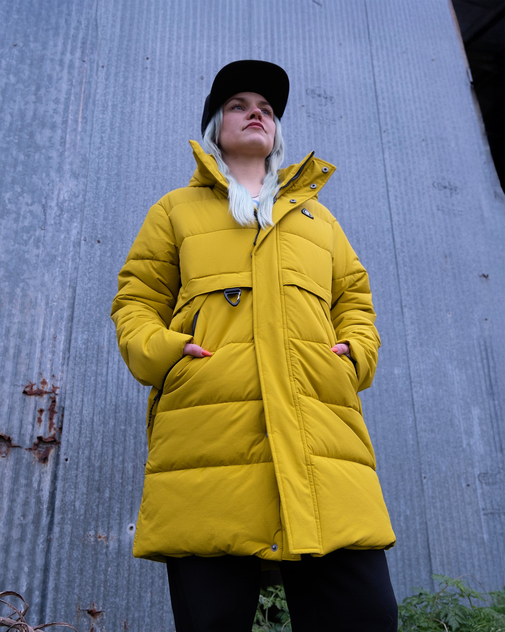 A person with long, light hair in the limited edition yellow beardedgoat NEW — MERIDIAN™ PARKA and a black cap stands before a corrugated metal wall, gazing upward with hands in their pockets.