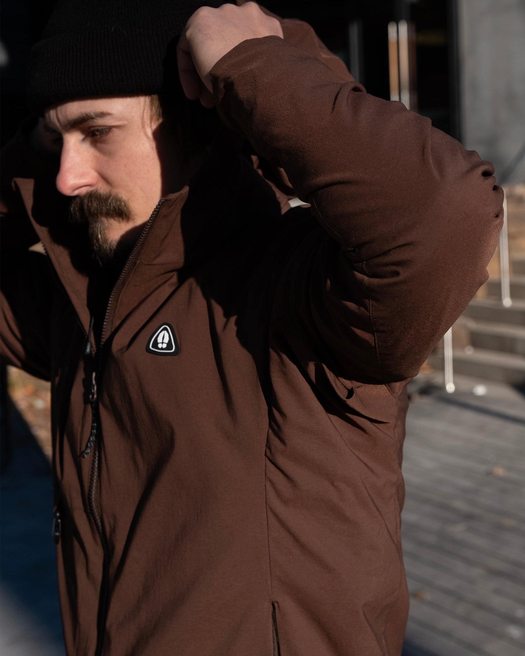 A mustached man in a black beanie adjusts his hood outdoors while wearing the NEW — OSLO Rover™ Performance All-Season Jacket from beardedgoat, with steps and a building entrance visible in the background.
