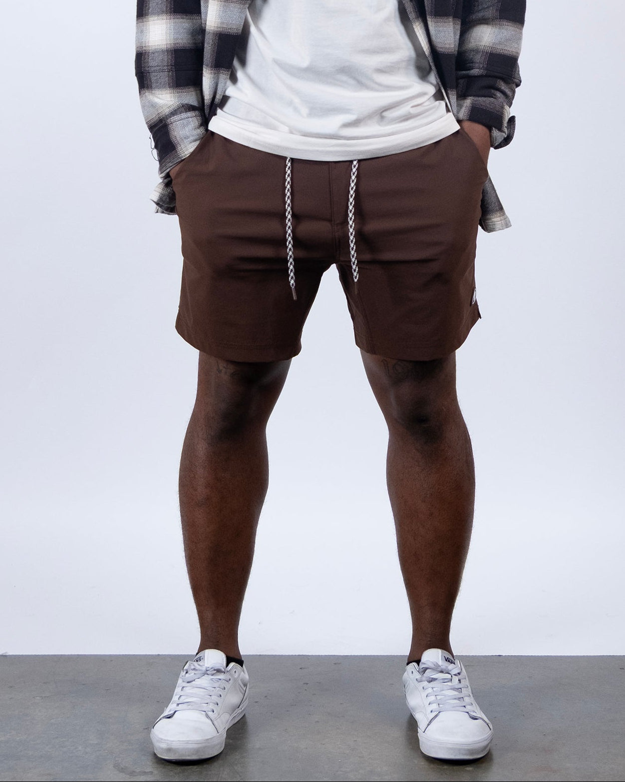A person stands with hands in pockets, wearing BEARDEDGOAT Men's Rover Hybrid Short in Terra Sienna, styled with a white drawstring, a white T-shirt, an open plaid shirt, and white sneakers against a plain light background.