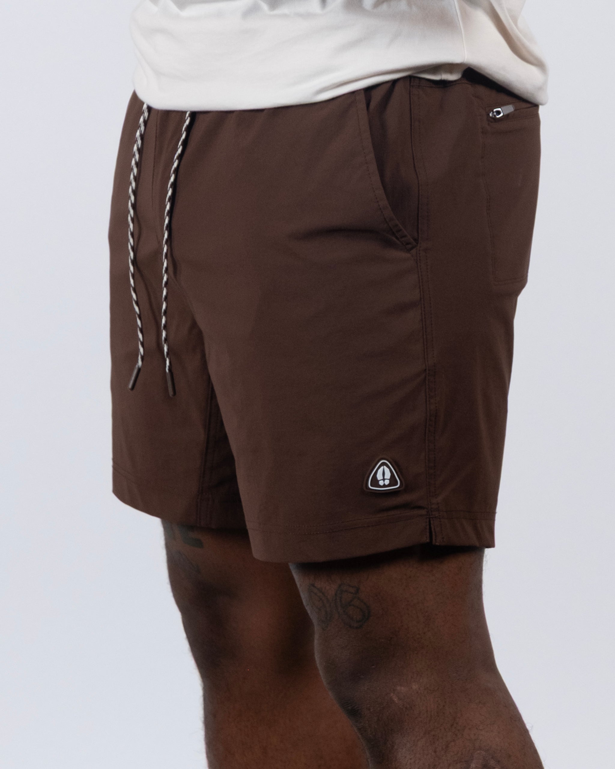 A person wears BEARDEDGOAT Men's Rover Hybrid Shorts in brown, featuring a small black and white logo on the leg, black and white drawstrings, and pockets. The above-the-knee shorts highlight thigh tattoos.