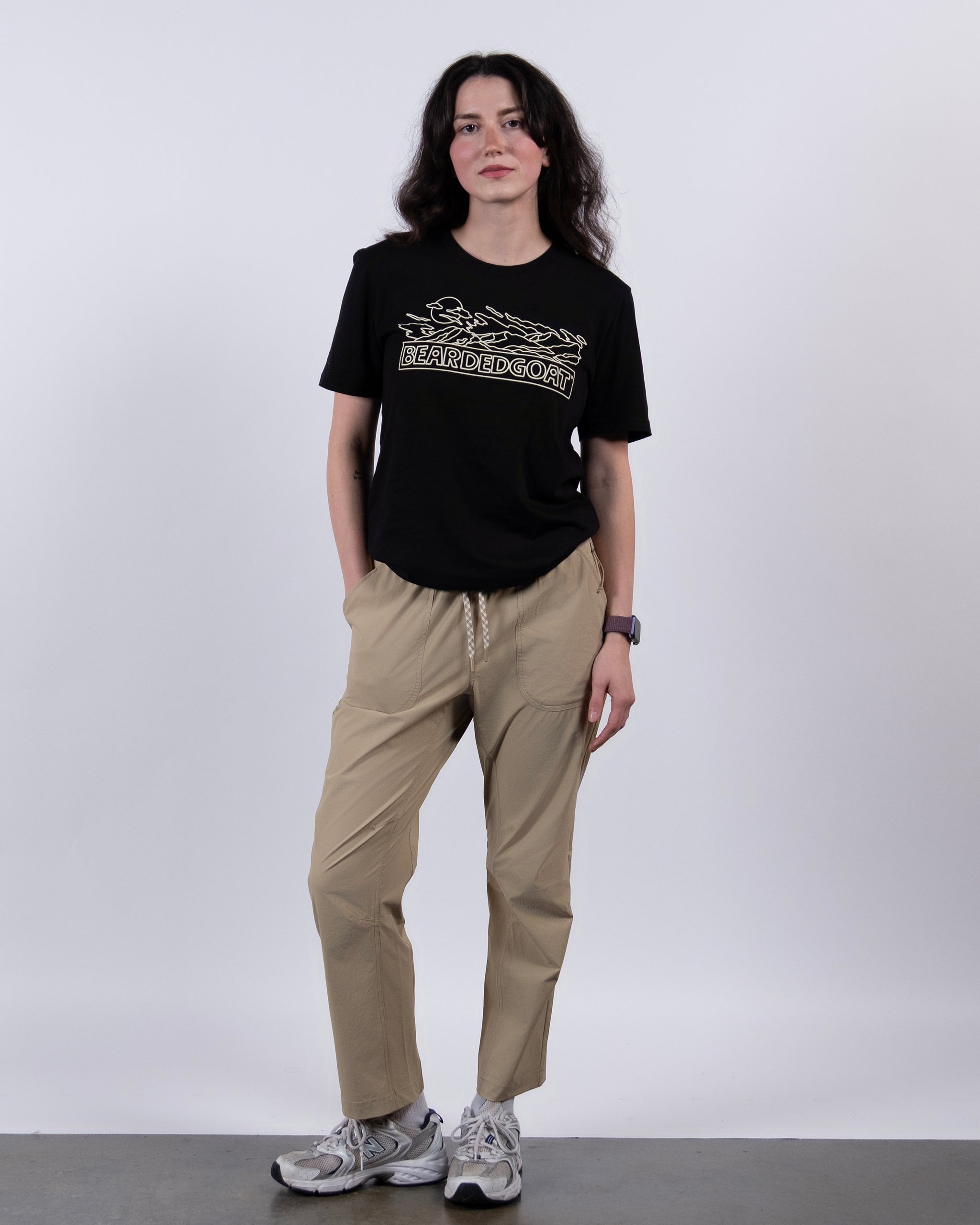 A woman stands against a white background wearing a black BEARDEDGOAT graphic t-shirt, NEW — Summit™ Pant in beige by BEARDEDGOAT, white sneakers, and a watch, with one hand in her pocket.