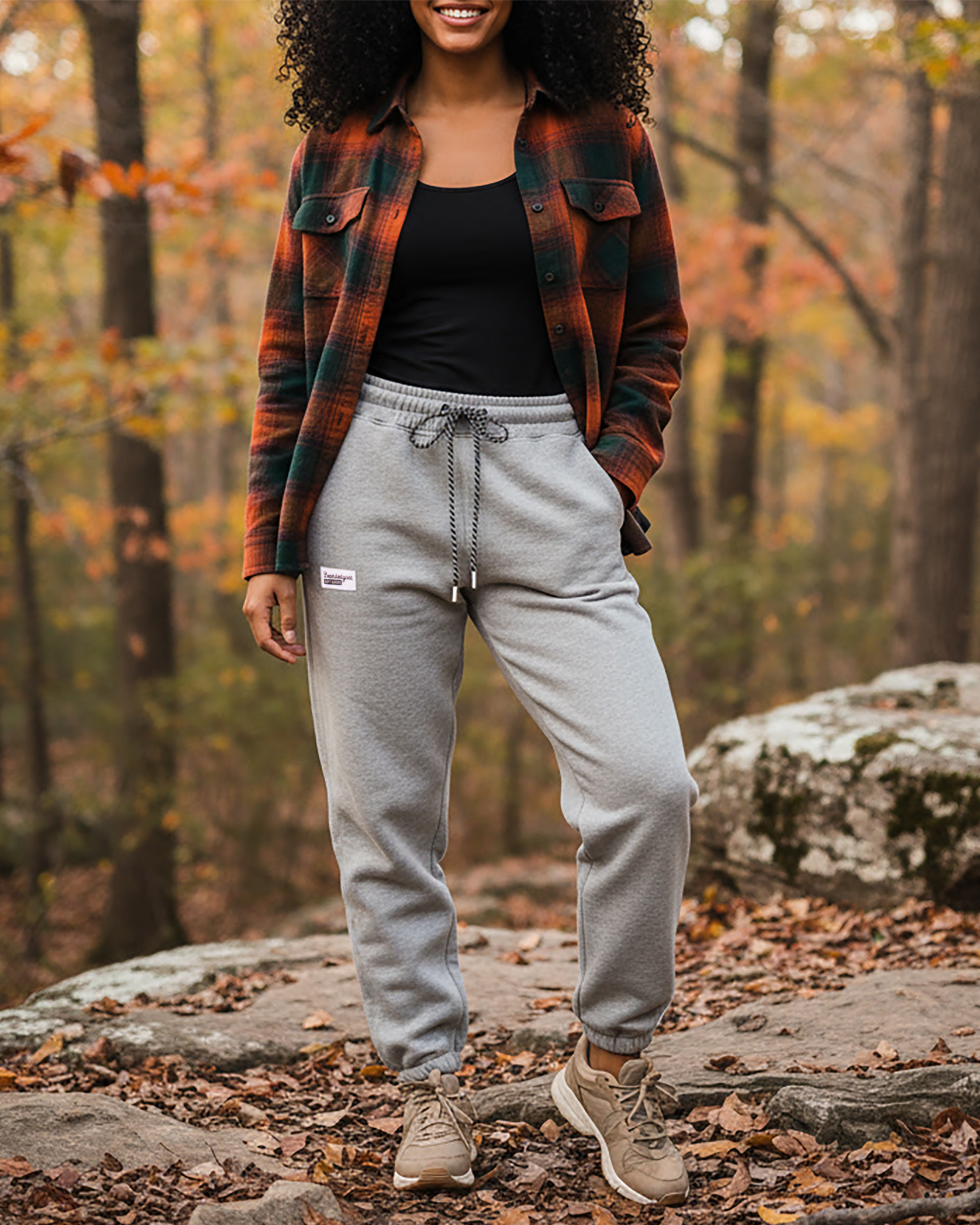 A person stands outdoors on a rocky path in the woods, wearing a green and orange plaid shirt over a black top, BEARDEDGOAT NEW — Overnight™ Classic Jogger sweatpants in heather grey, and tan sneakers amid autumn trees.