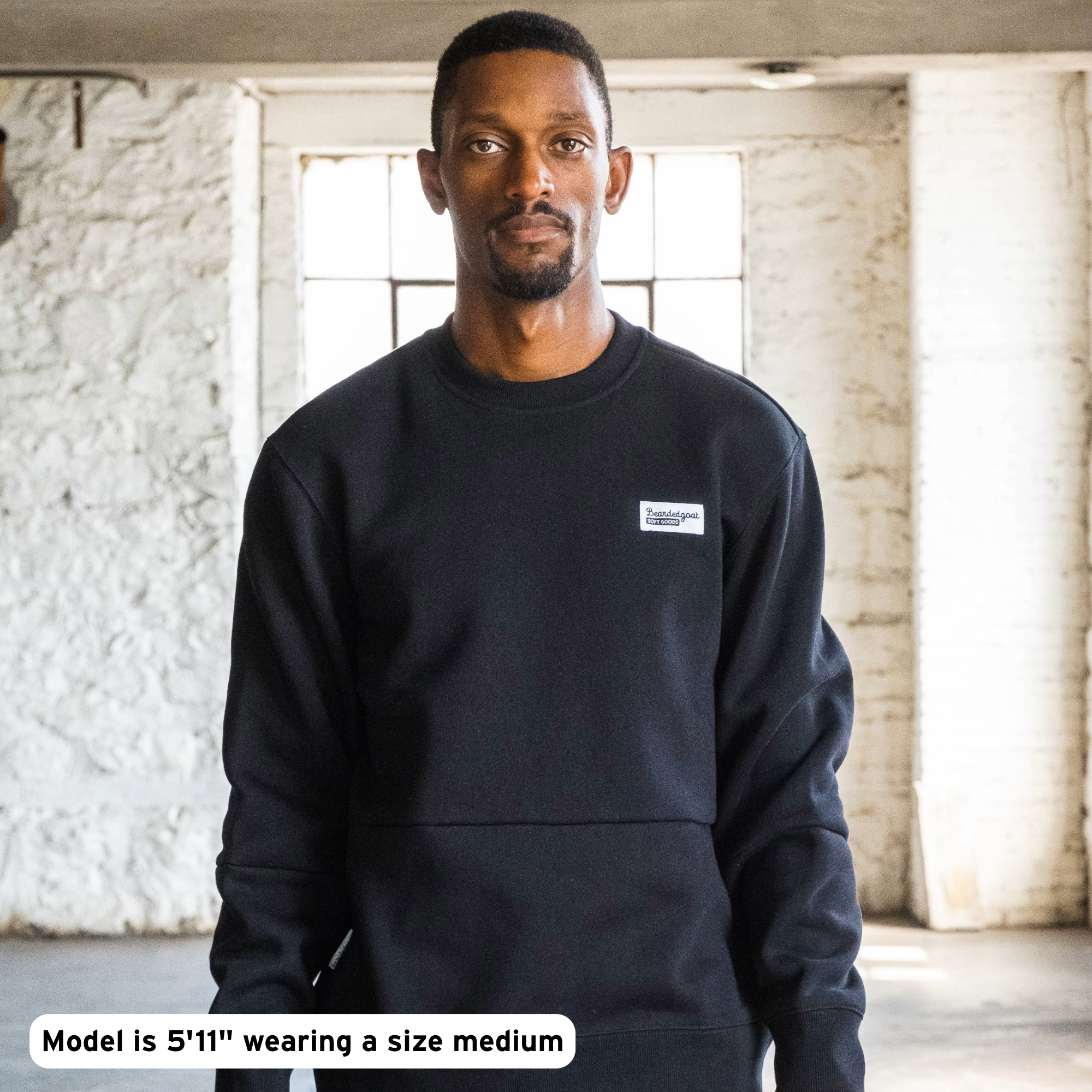 A man stands indoors wearing the beardedgoat Men's Void Crew in black, featuring a small chest logo. White brick walls and large windows form the backdrop. Text reads: Model is 5'11" and wears size medium.